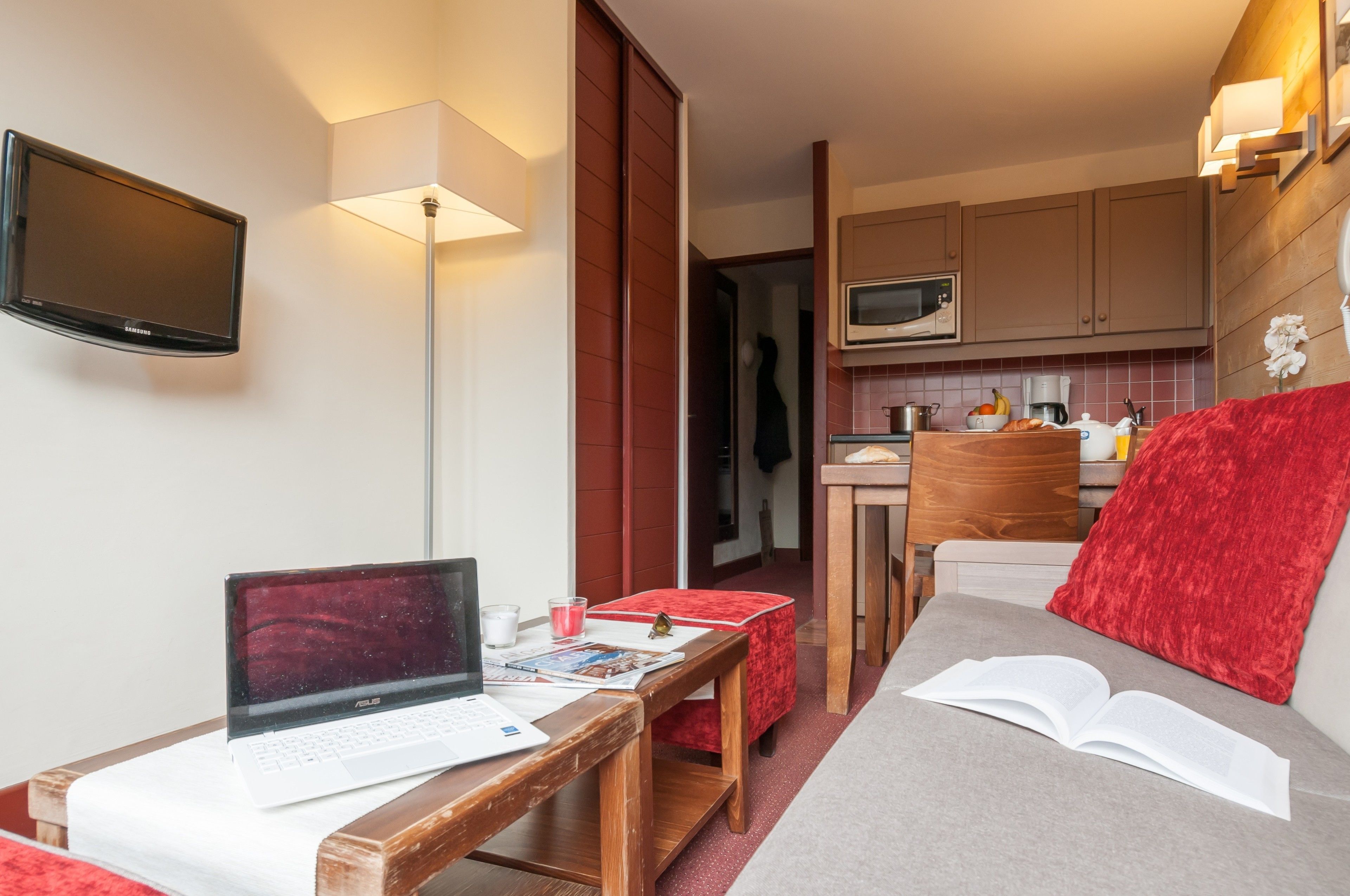 A compact living space with a sofa, a small table with a laptop, a flat-screen TV, and a kitchenette.