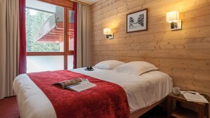 Habitación de hotel con pared de madera, cama con ropa blanca, manta roja, mesita de noche con lámpara y ventana con cortinas.