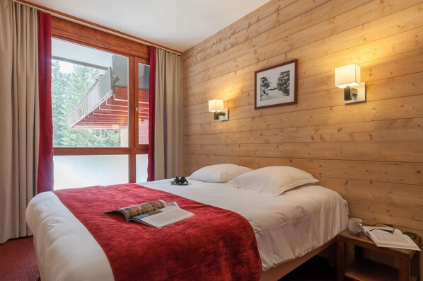 Hotellihuone, jossa on puinen seinä, valkoisilla lakanoiden peitetty sänky, punainen heittopeitto, yöpöytä lampun kanssa ja ikkuna, jossa on verhot.