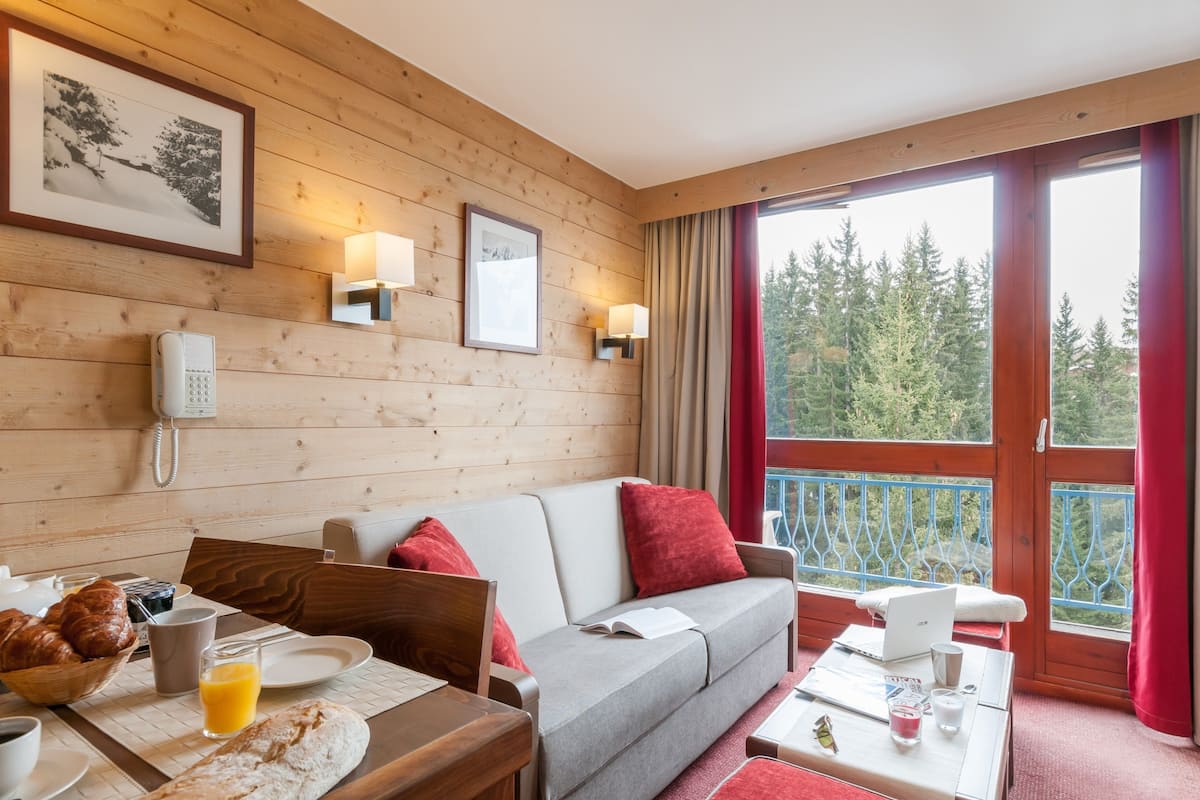 A cozy room with a wooden wall, a sofa, a dining table with breakfast items, and a balcony with a view of trees.