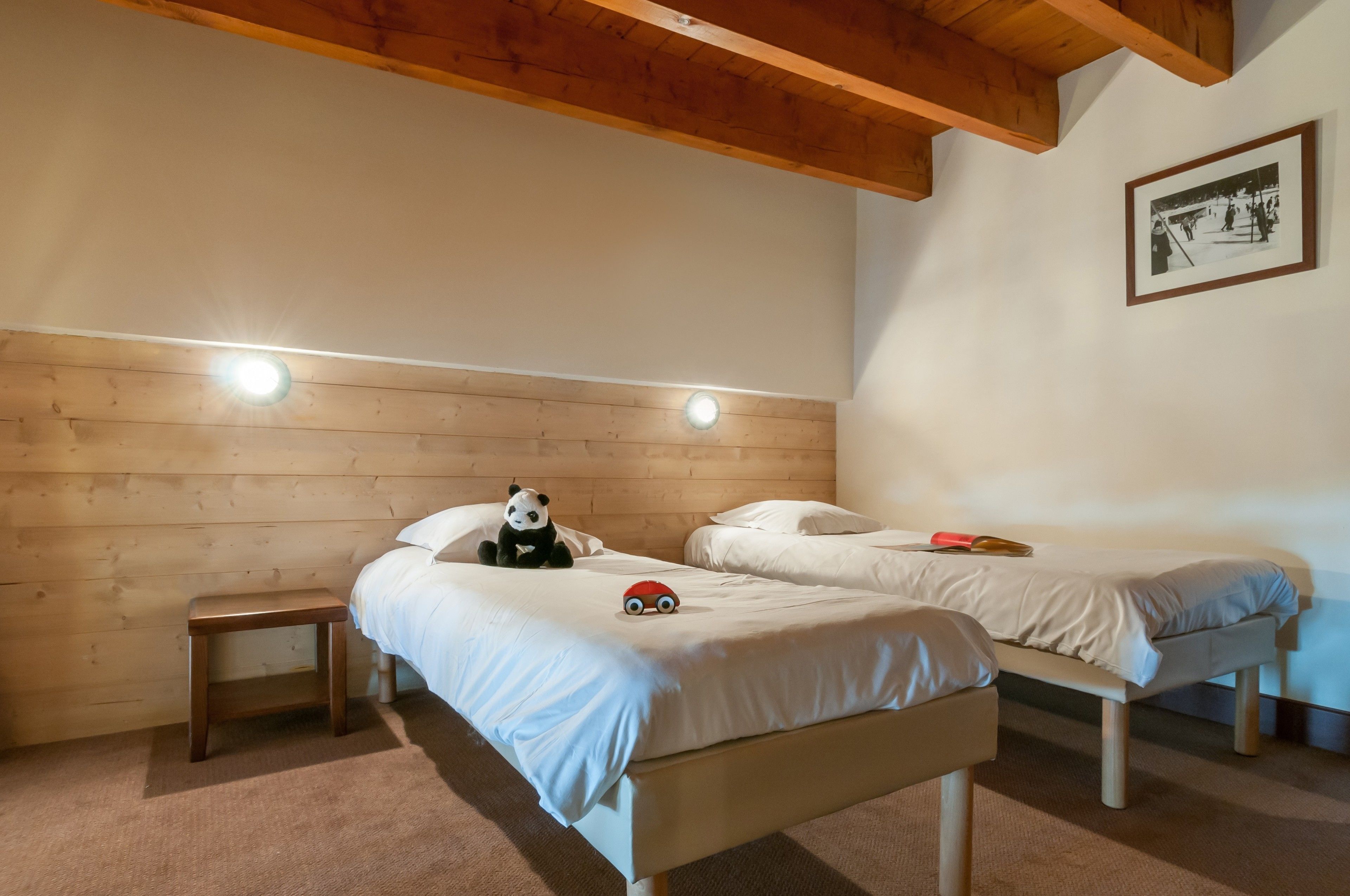 Quarto com duas camas, uma cabeceira de madeira, uma mesa pequena e uma fotografia emoldurada na parede.