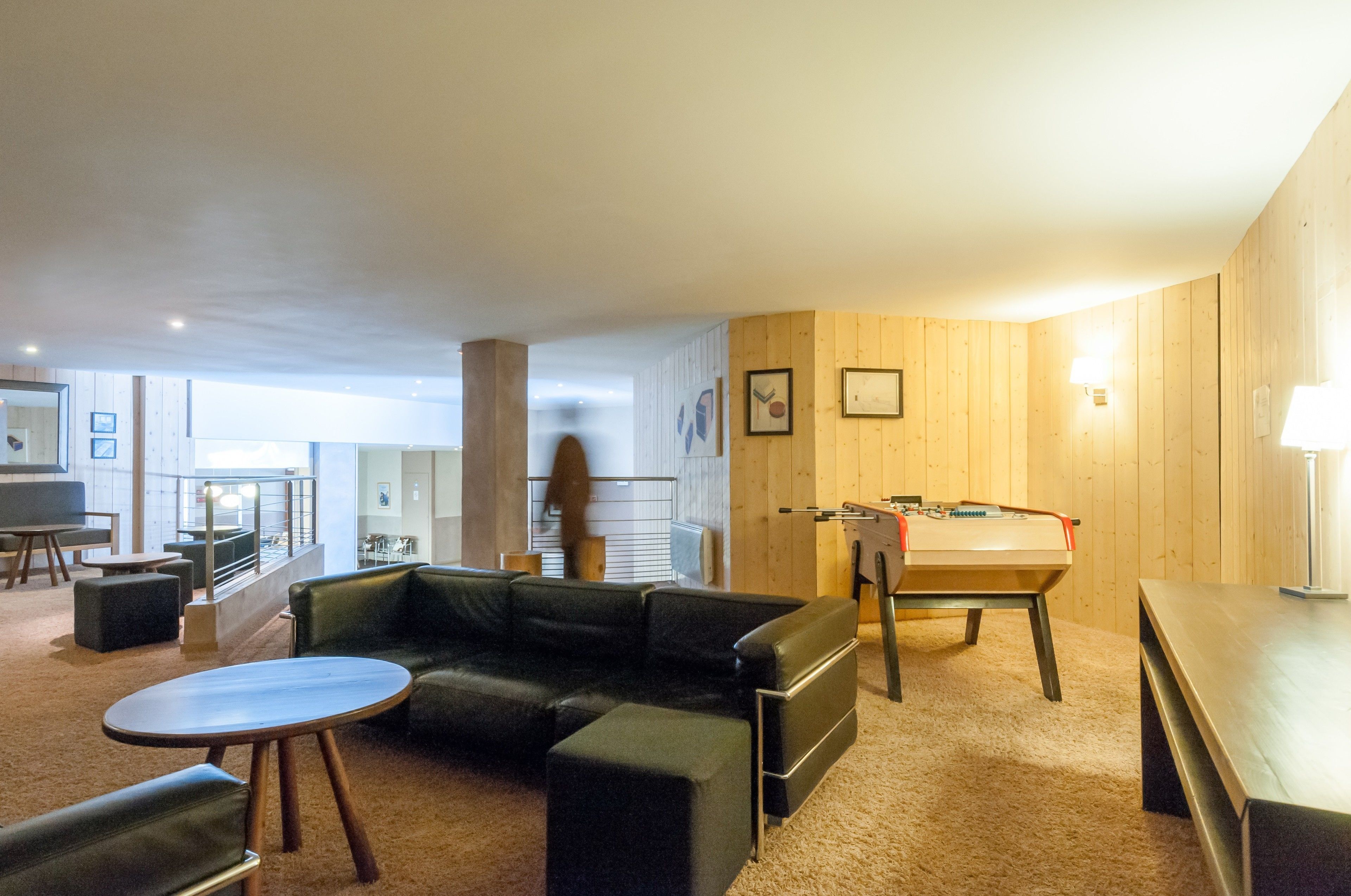 A modern living room with a sofa, a coffee table, and a foosball table. There is a balcony with a railing and a view of the outside.