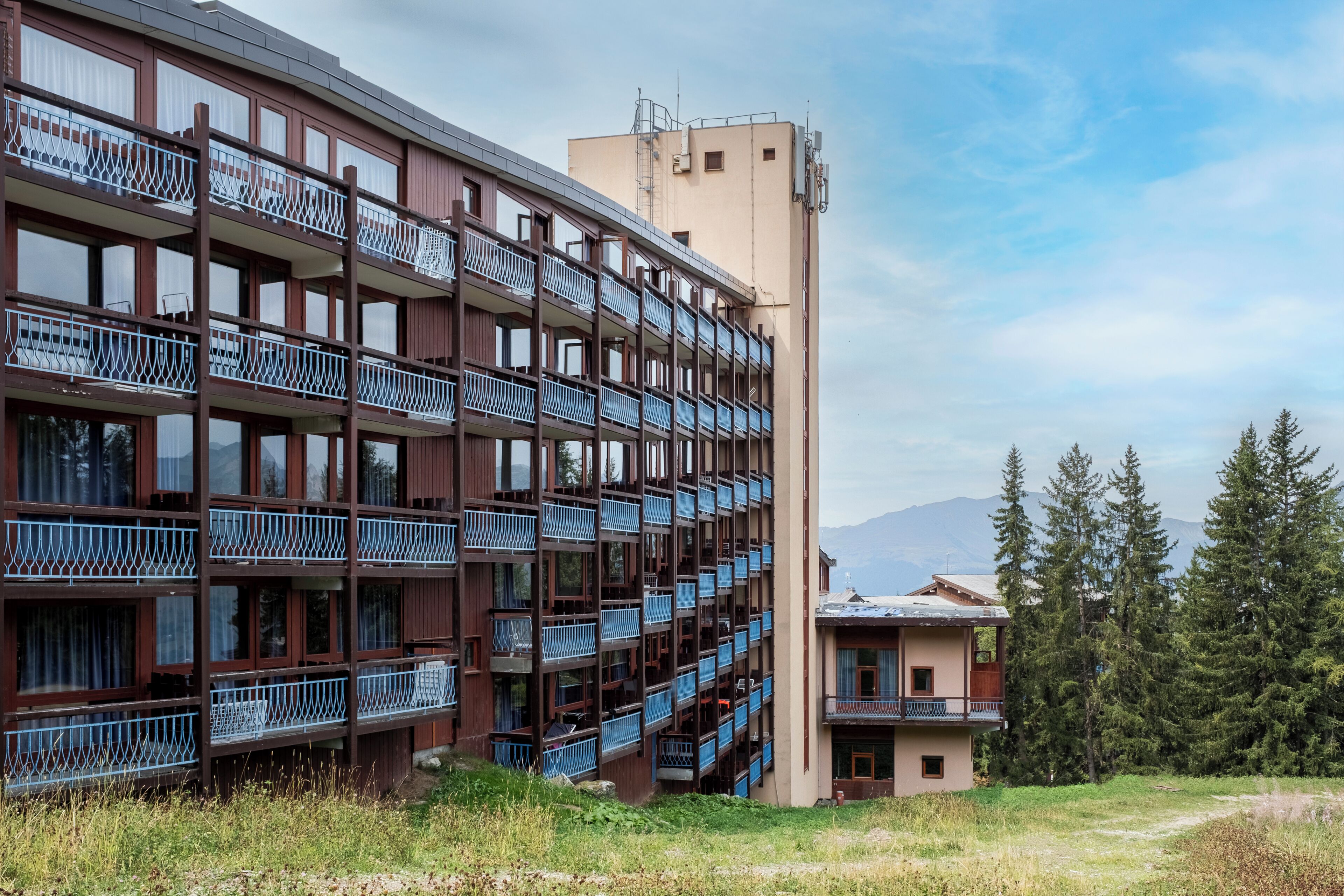 Ein mehrstöckiges Gebäude mit Balkonen, ein separates Eingangsbau und eine gebirgige Kulisse.