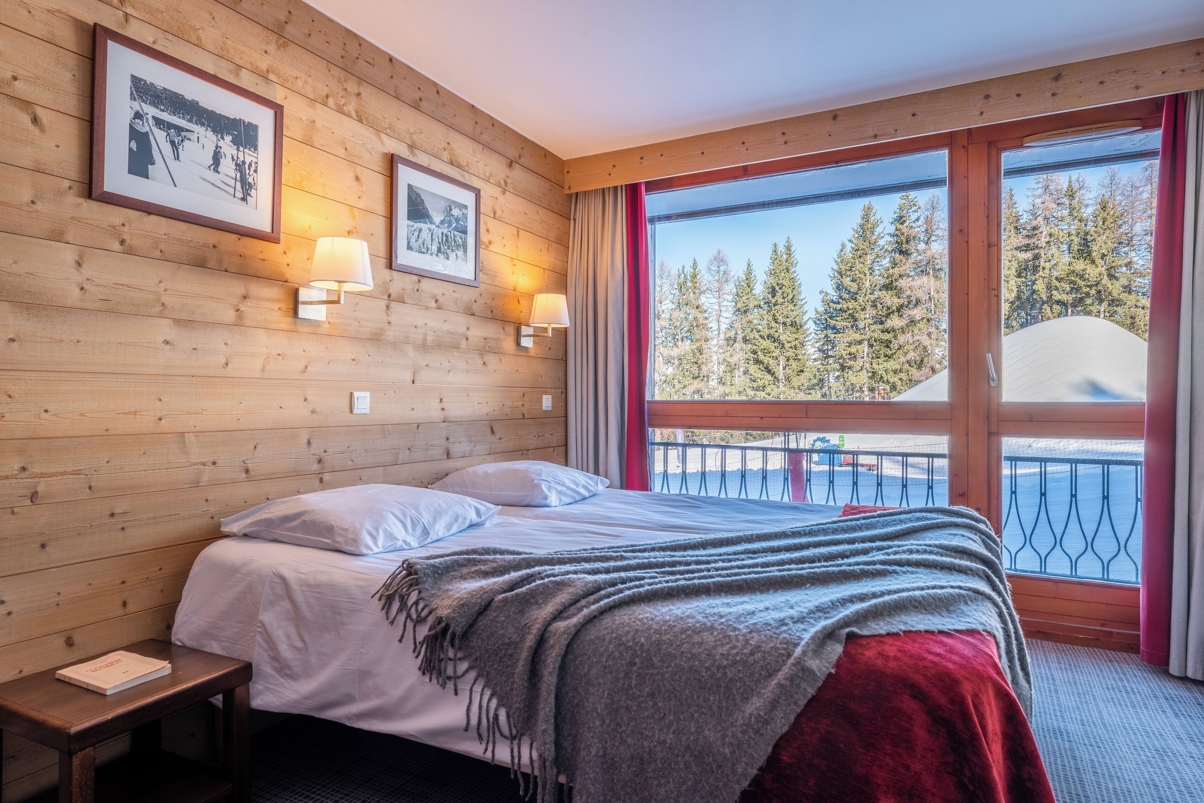 Ein Zimmer mit Holzwänden, einem Bett, zwei Wandlampen und einem großen Fenster mit Blick auf Bäume und ein Boot.