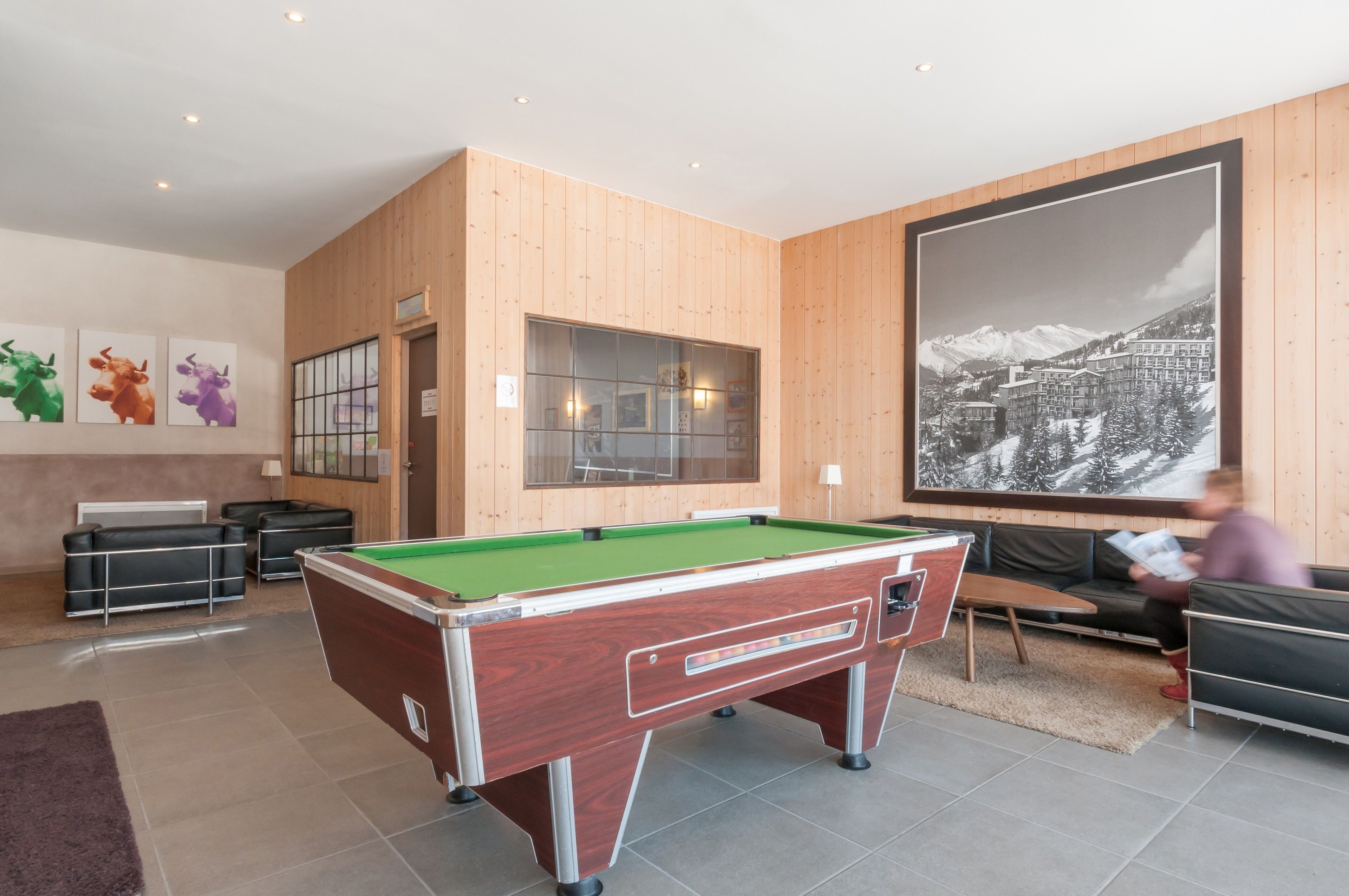 Una mesa de billar en una habitación con paneles de madera, un cuadro grande enmarcado con un paisaje montañoso y un sofá.