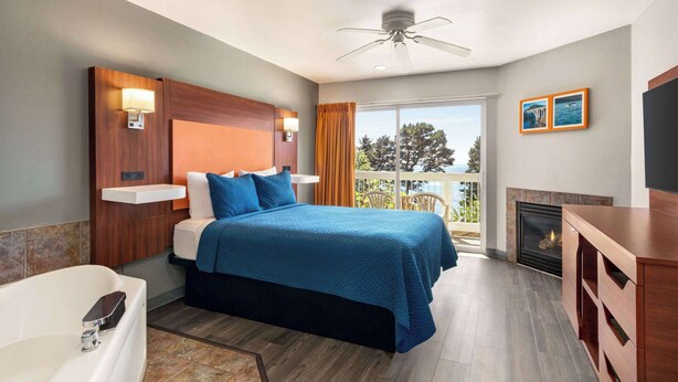 A hotel room with a bed, a ceiling fan, a fireplace, a balcony with a view, and a TV. - Travelodge by Wyndham Depoe Bay (Depoe Bay)