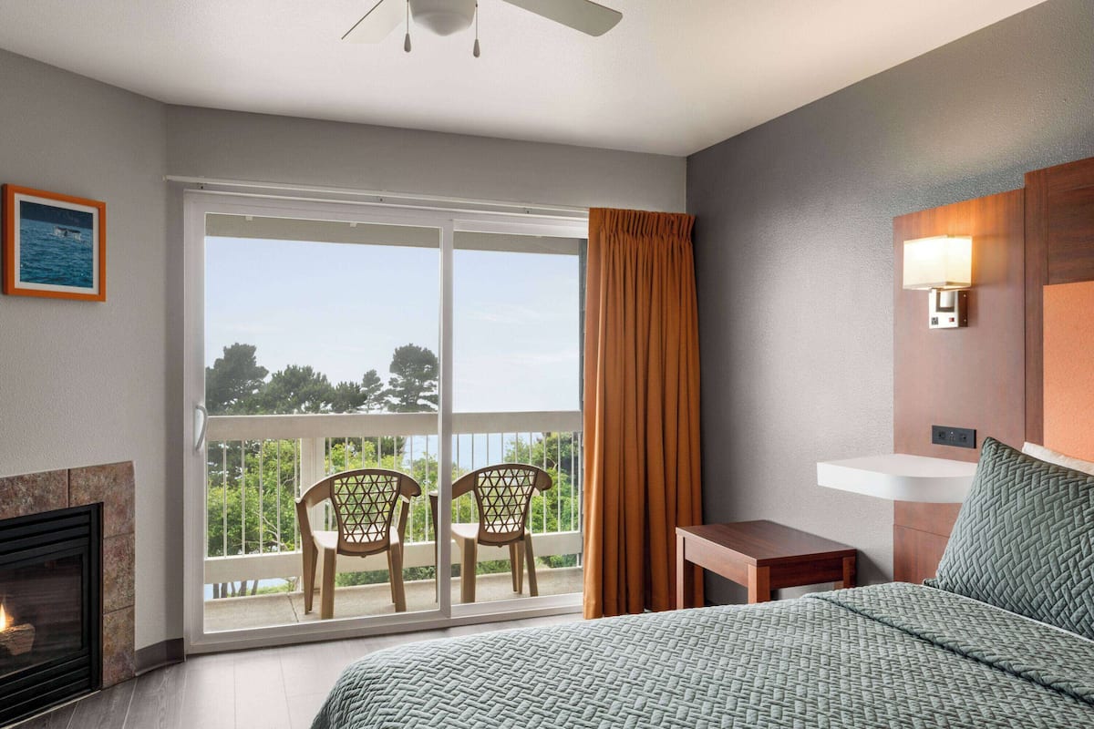 Habitación de hotel con cama, chimenea, balcón con sillas y vista al exterior.