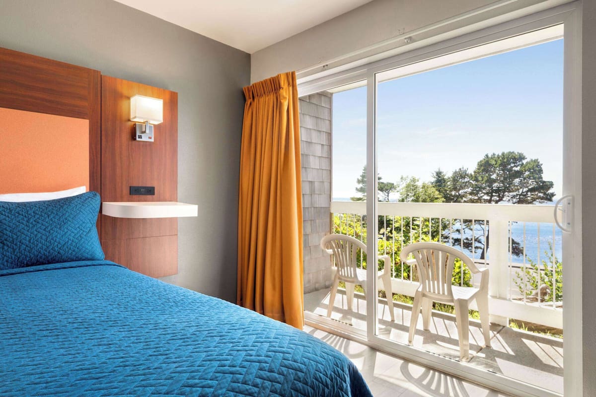 Habitación de hotel con cama, mesita de noche, lámpara de pared, balcón con sillas y vistas al exterior.
