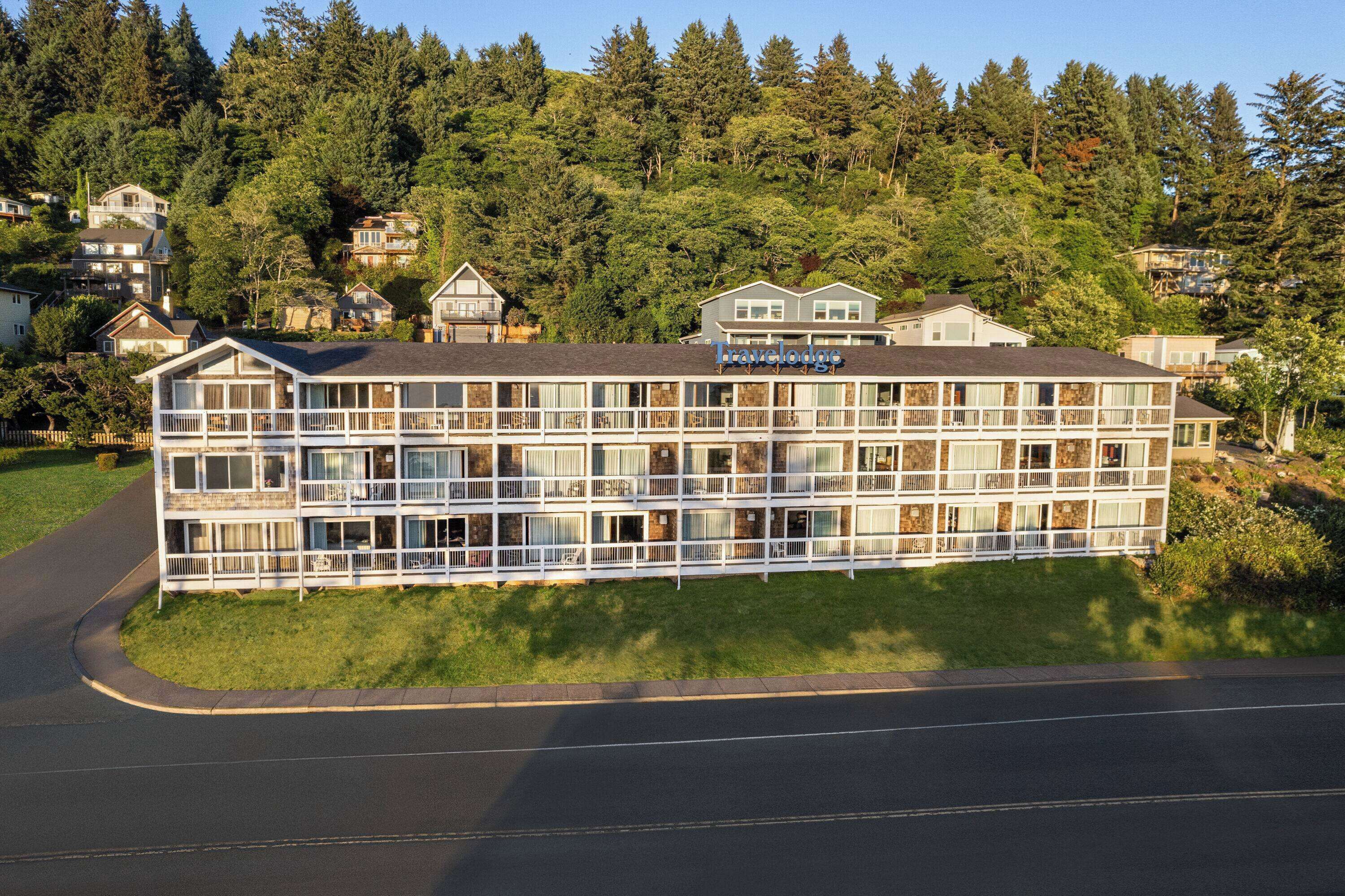 Un hotel de varios pisos con balcones, situado en una zona residencial con casas y árboles al fondo.