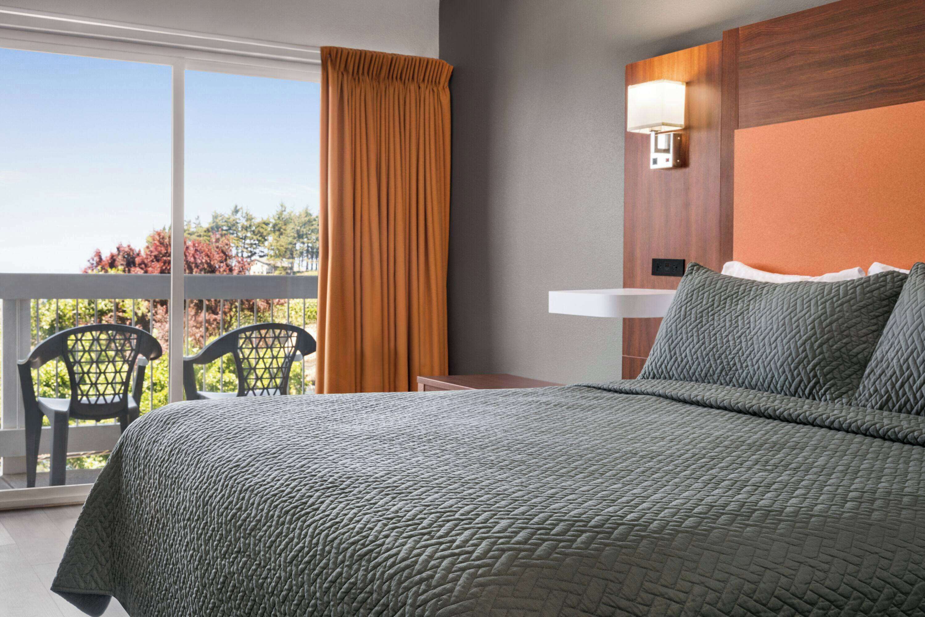 A hotel room with a bed, a nightstand, a lamp, a window with curtains, and a balcony with chairs.