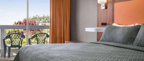 A hotel room with a bed, a nightstand, a lamp, a window with curtains, and a balcony with chairs.