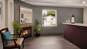 A living room with a fireplace, a large mirror, a window with a view of a house, and a wooden reception desk. - Travelodge by Wyndham Depoe Bay (Depoe Bay)