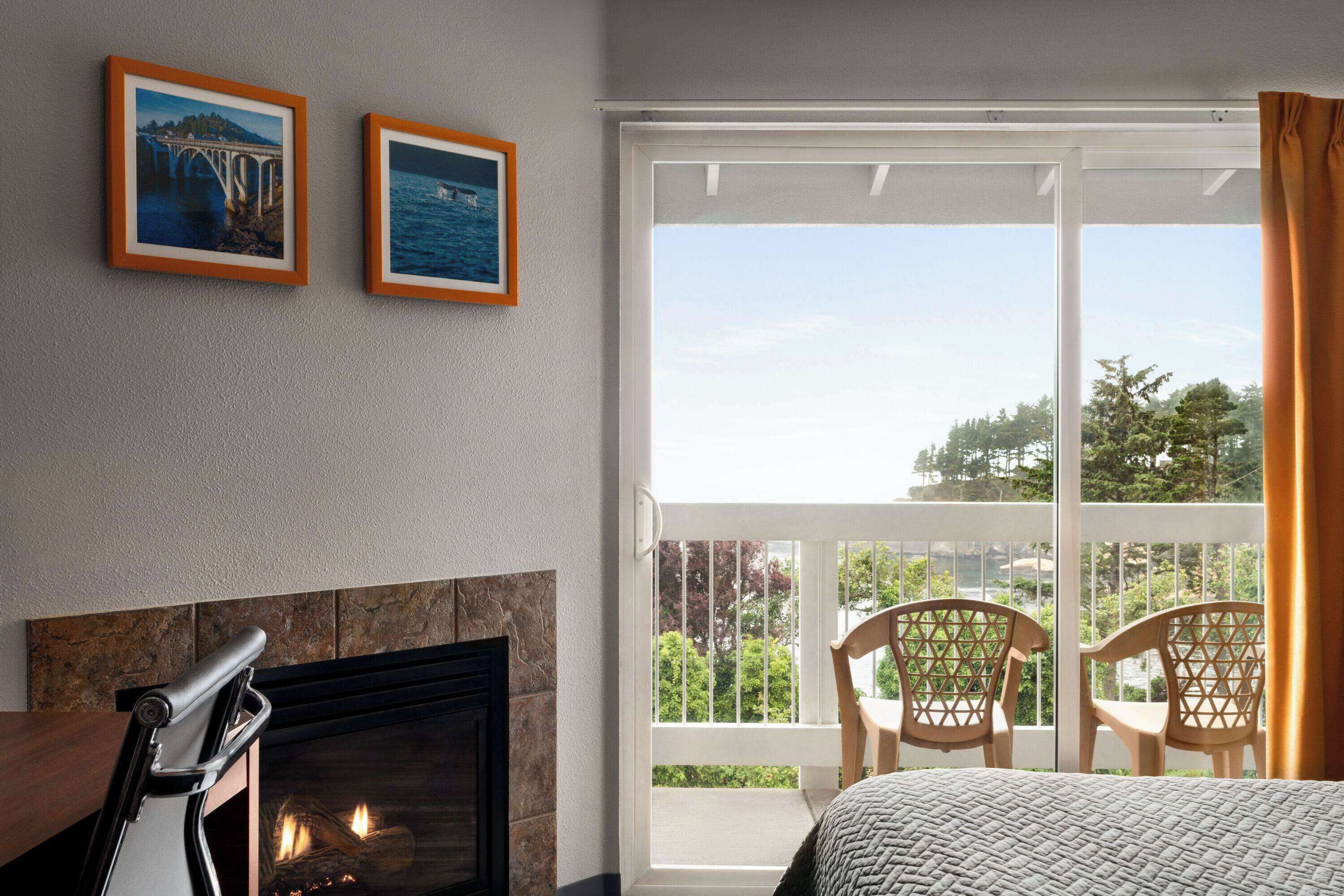 A room with a fireplace, two framed pictures on the wall, a balcony with chairs, and a view of greenery.