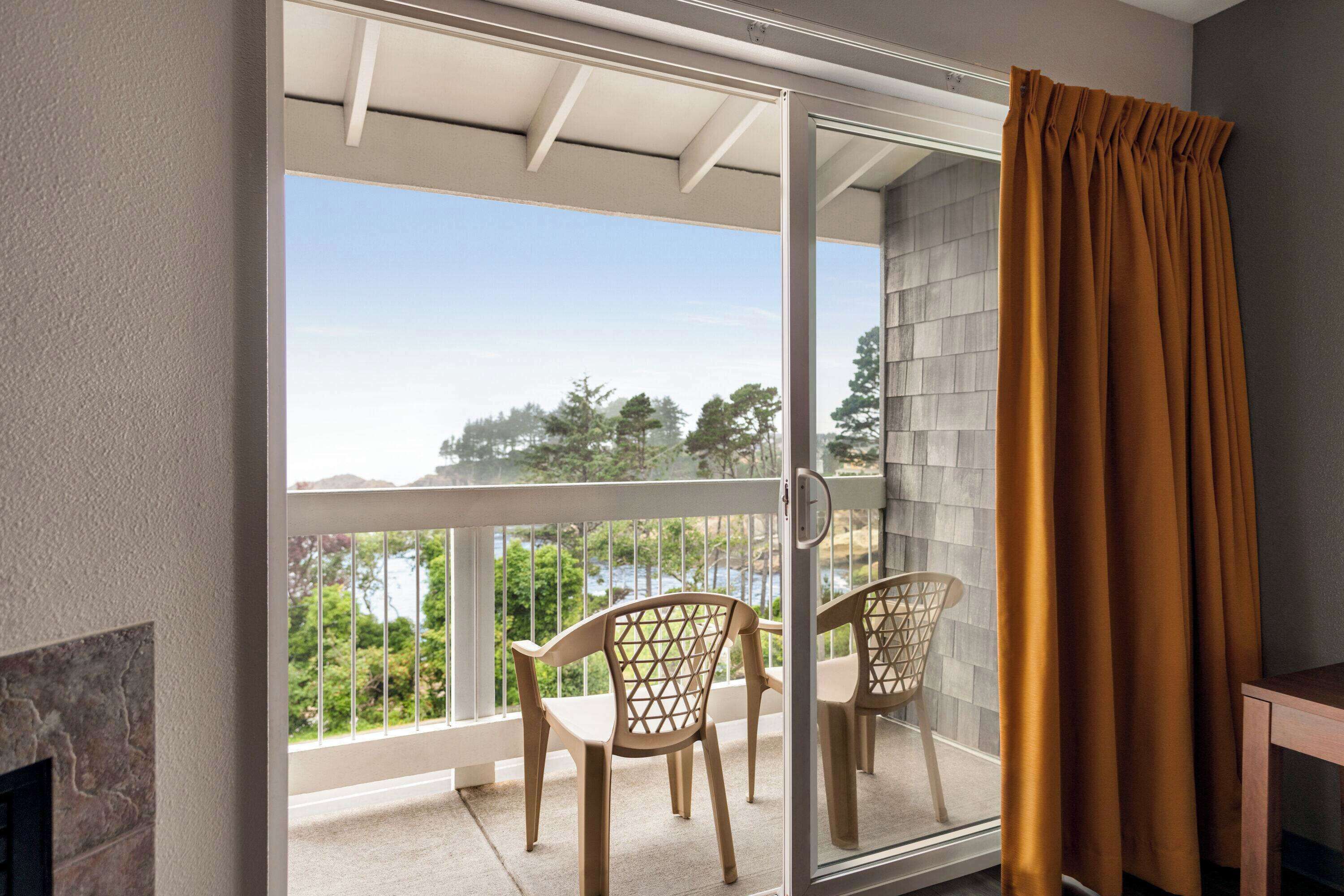 Ein Balkon mit Blick auf Bäume und ein Gewässer, eine Schiebeglastür sowie zwei Korbstühle.