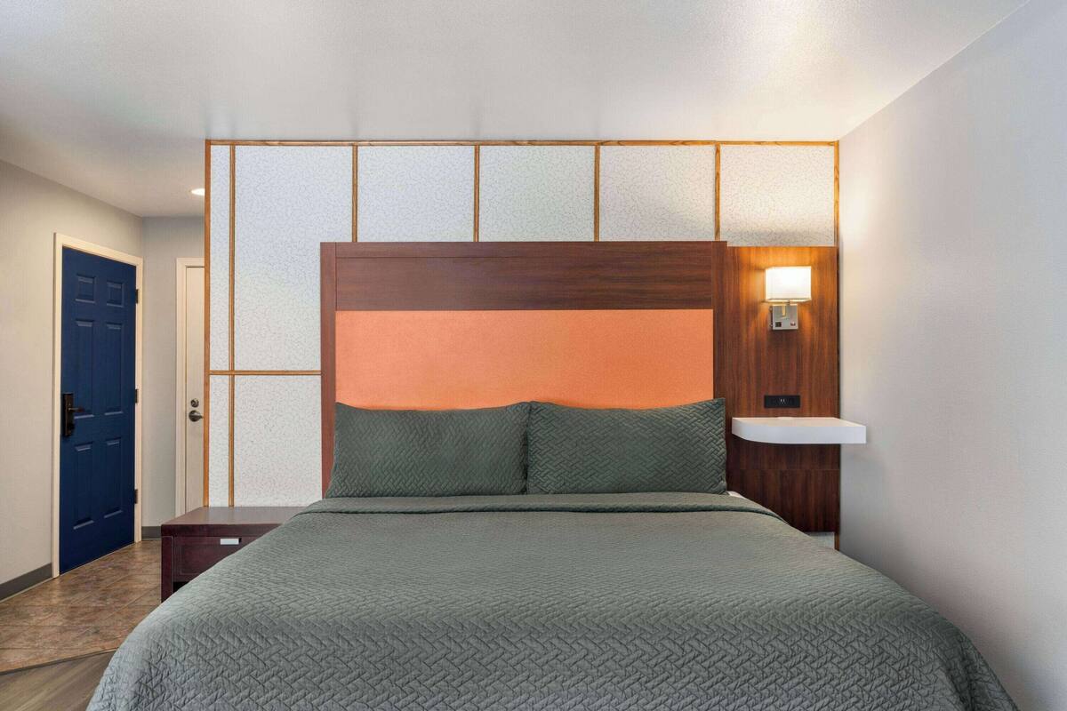 Una habitación de hotel con una cama, una mesita de noche, una lámpara de pared y una puerta.