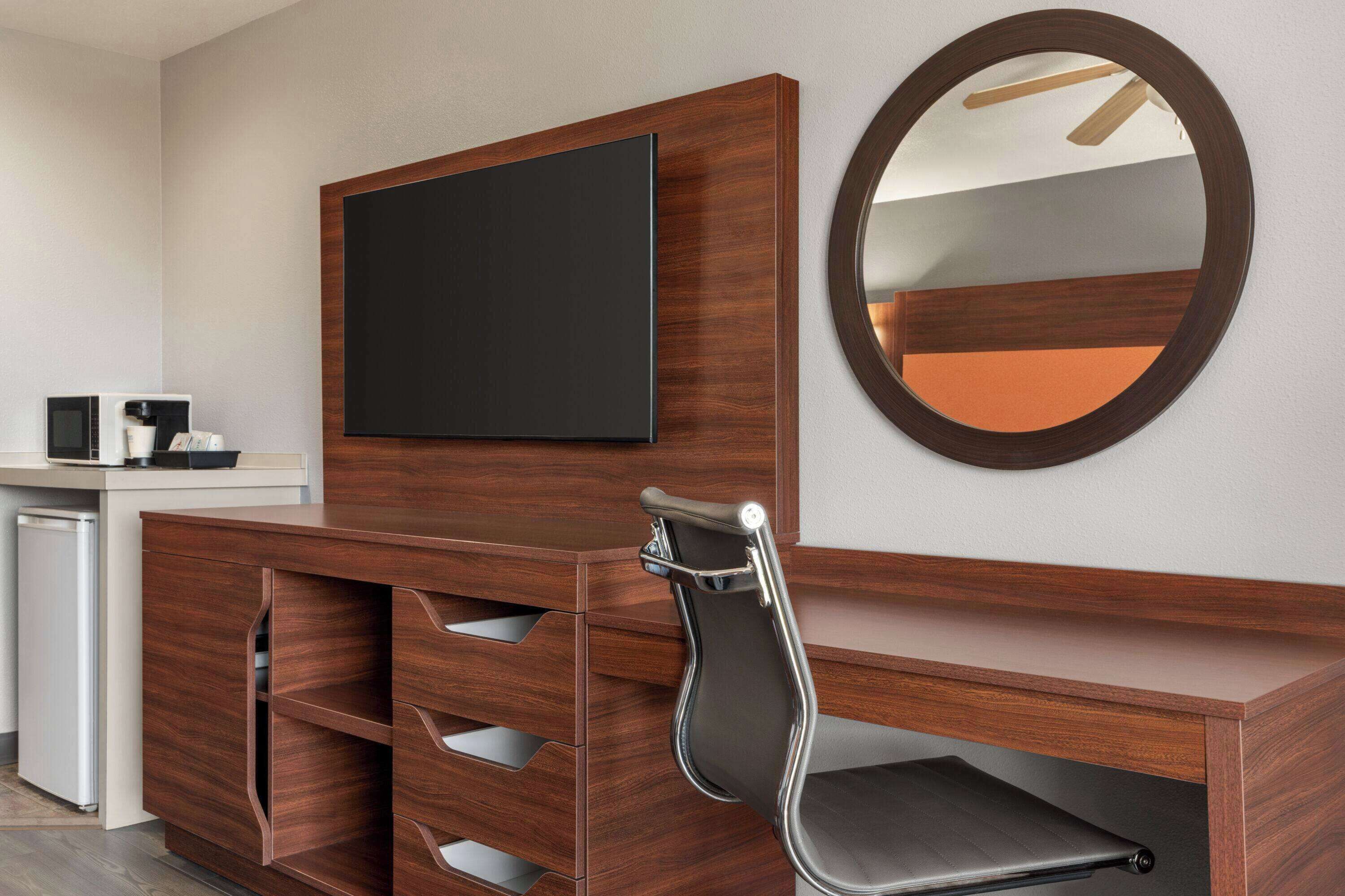 Une chambre d’hôtel avec un bureau en bois, un téléviseur à écran plat, un miroir rond et une chaise.