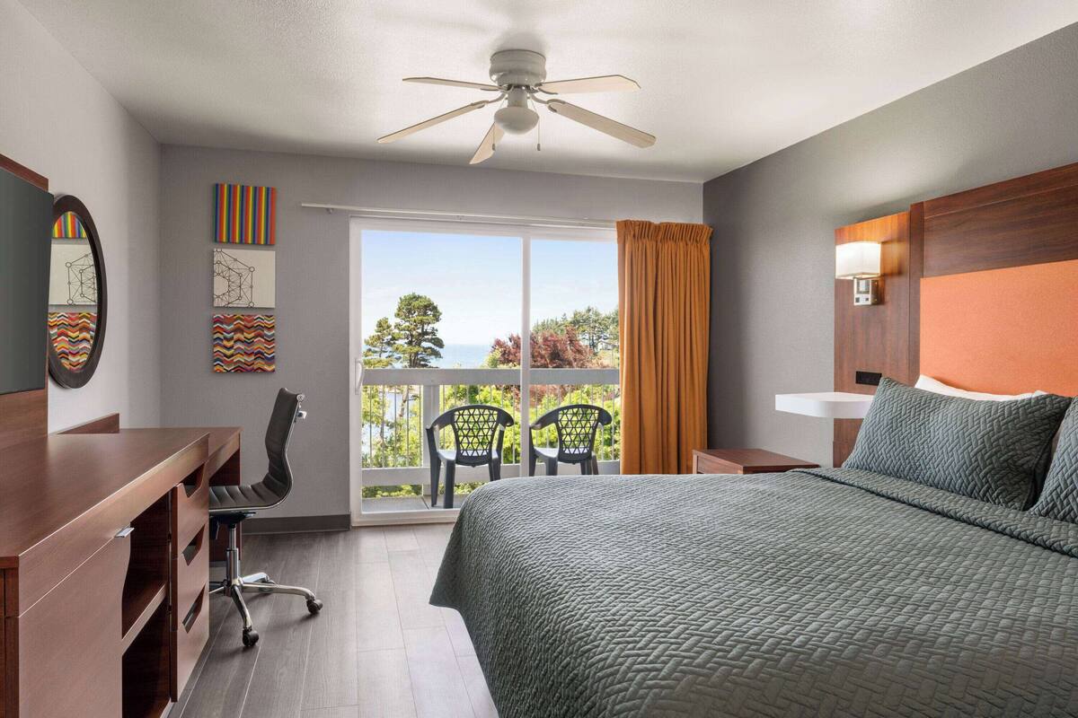 Habitación de hotel con cama, escritorio, sillas, ventilador de techo y balcón con vistas.