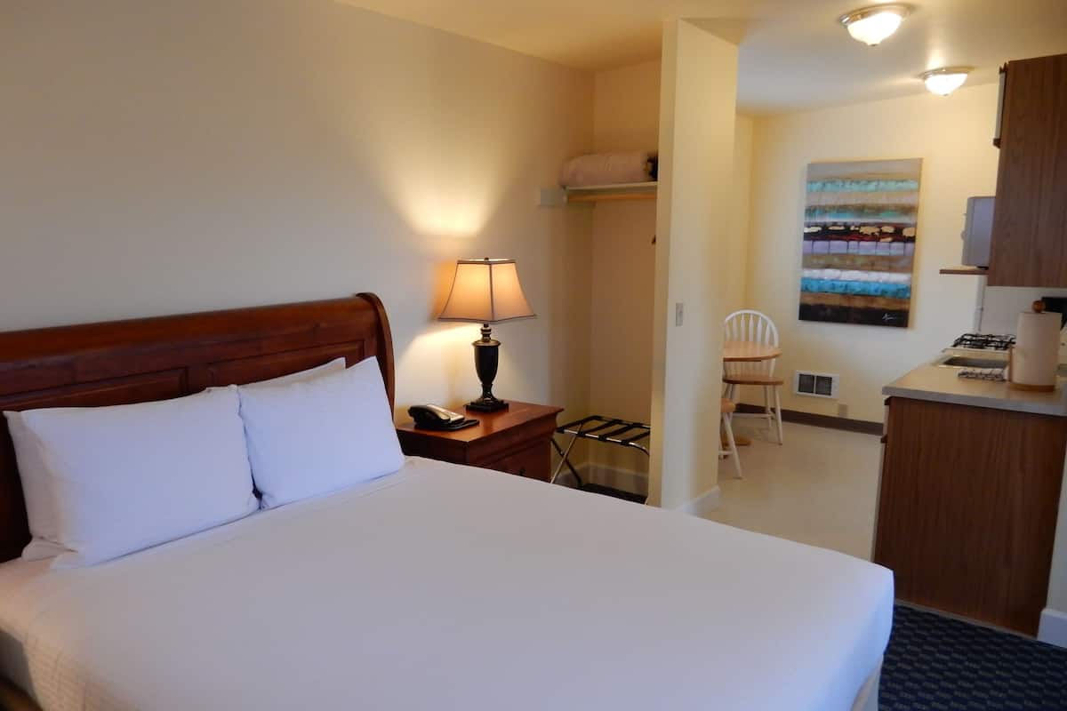 A hotel room with a bed, a bedside table with a lamp, a chair, a small kitchenette, and a painting on the wall.