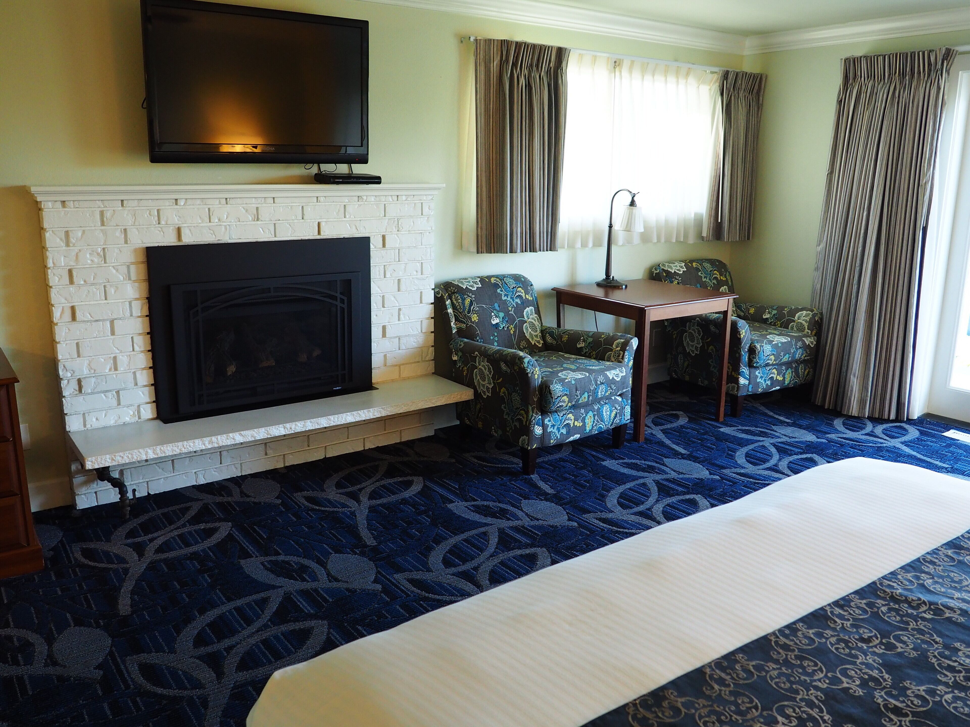 A hotel room with a fireplace, two patterned armchairs, a small table, and a large bed.