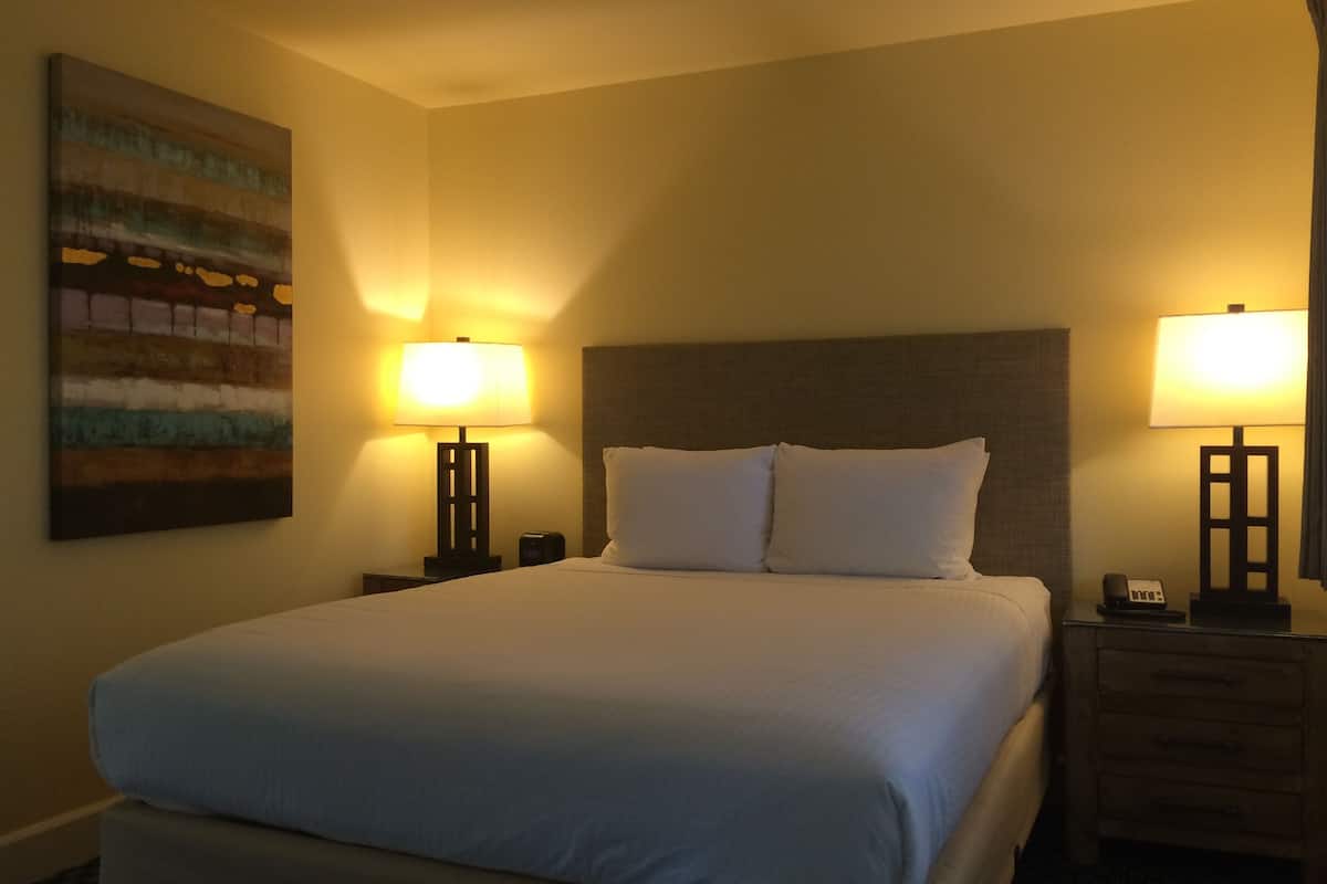 A hotel room with a large bed, two bedside tables, a nightstand, a wall-mounted painting, and two lamps.