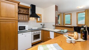 A kitchen with wooden cabinets, a microwave, an oven, a sink, and a window with curtains. - Madame Vacances Les Chalets des Alpages (La Plagne-Tarentaise)