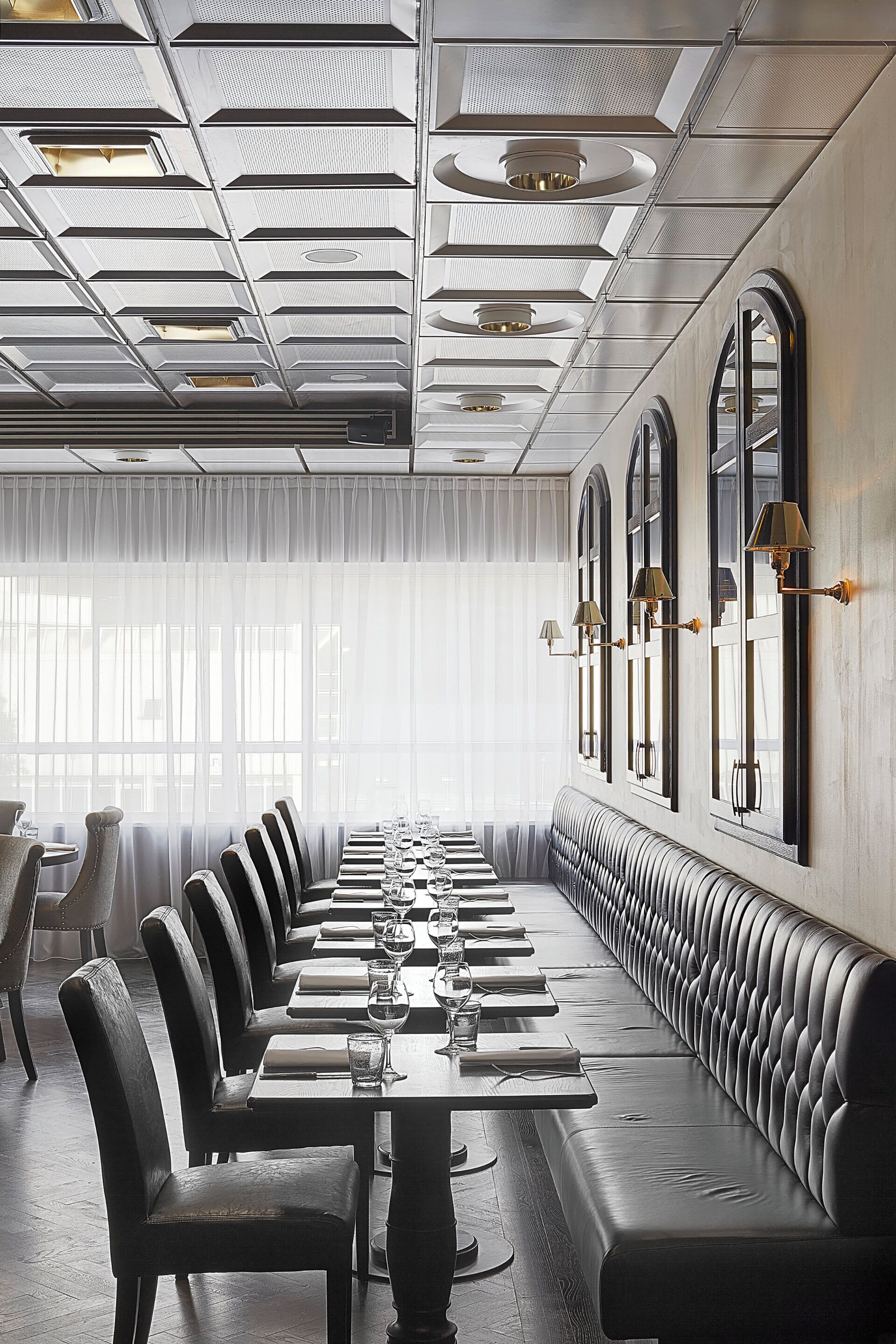 A long dining table with chairs and a bench, set against a wall with arched windows and wall sconces.