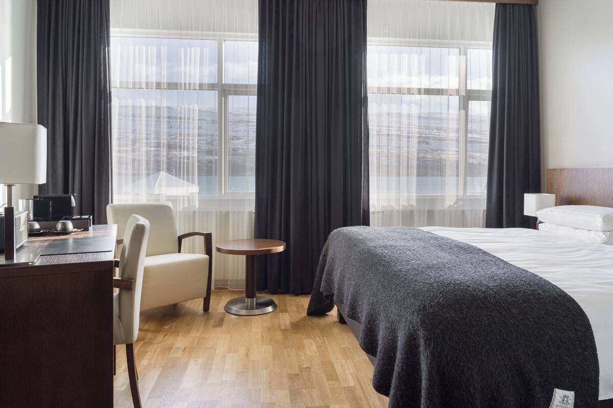 Ein Hotelzimmer mit einem Bett, einem Schreibtisch mit Lampe, einem Sessel, einem kleinen Tisch und großen Fenstern mit Vorhängen.