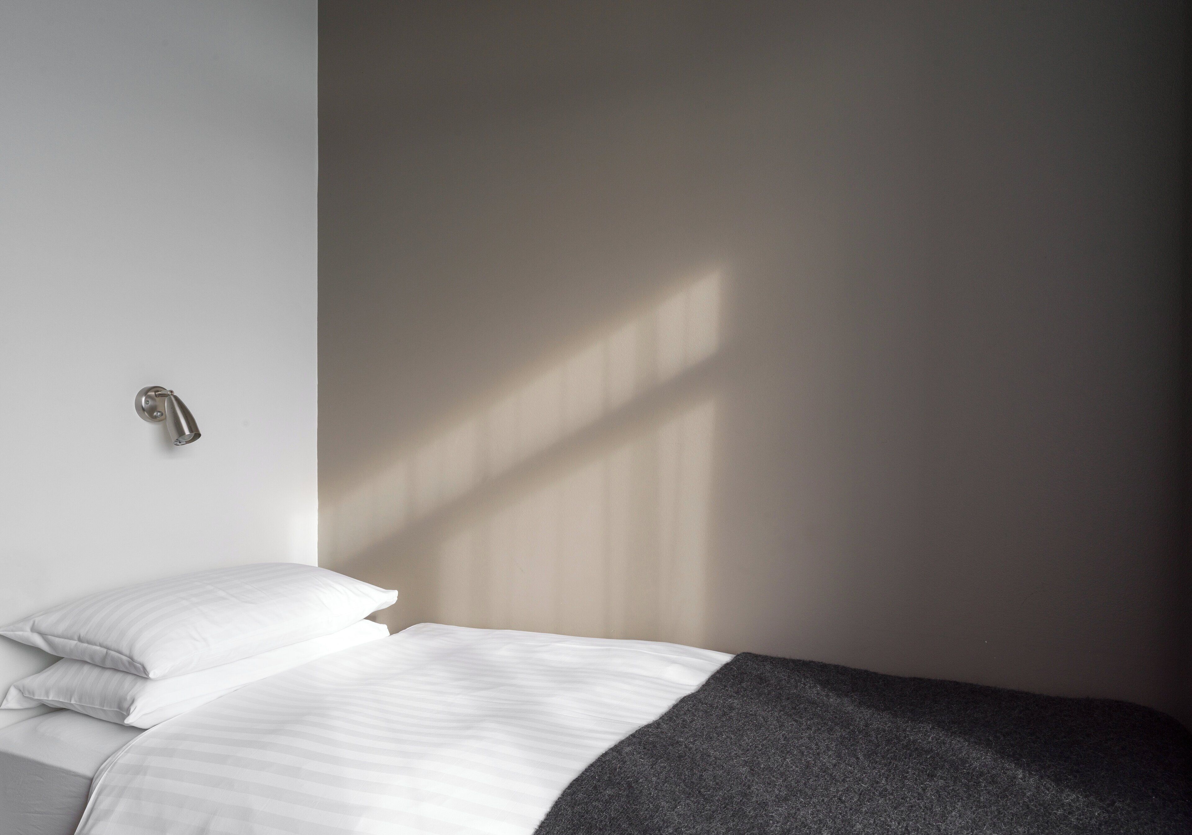 A minimalist bedroom with a bed, a wall-mounted light, and a window casting shadows.