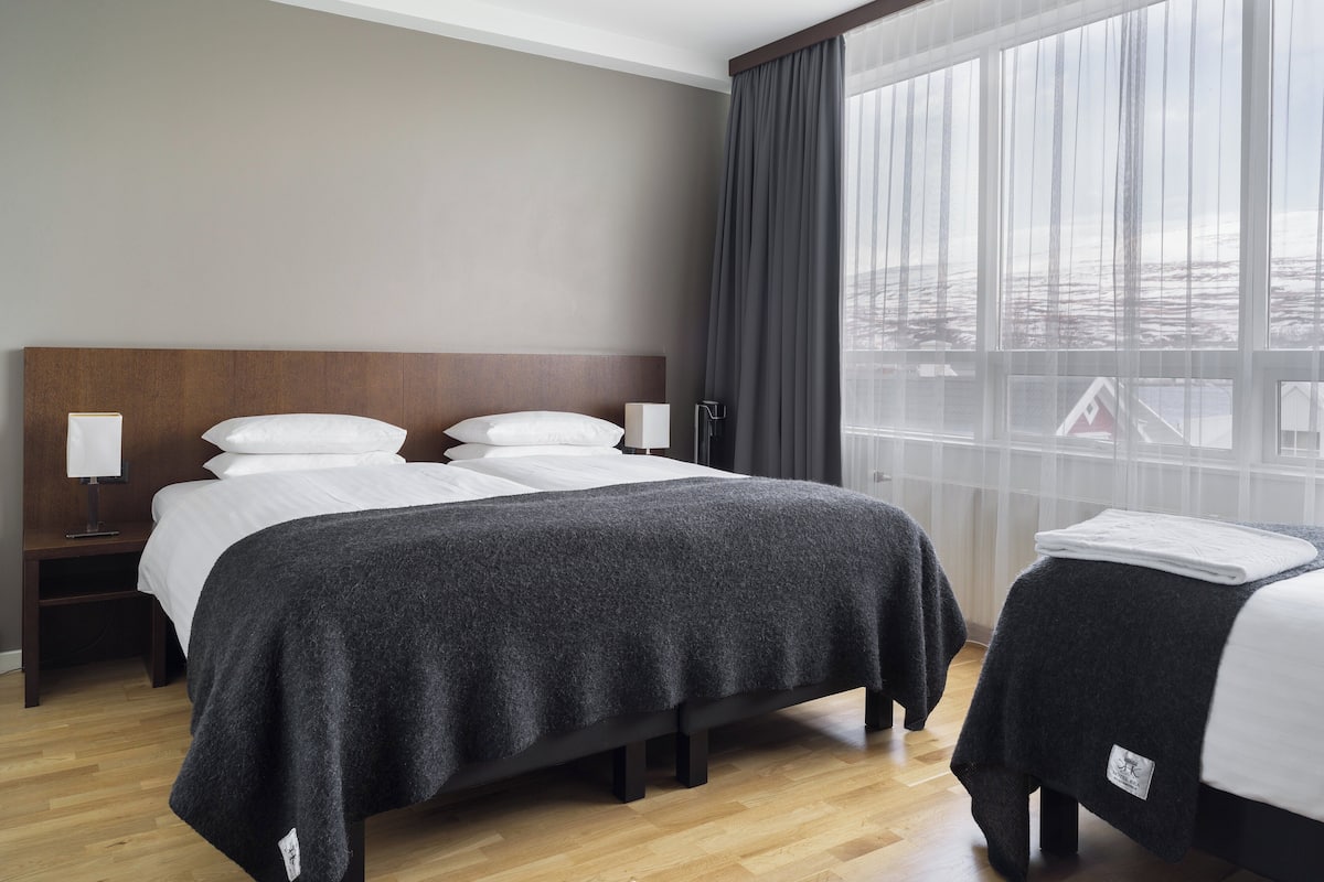 Ein Hotelzimmer mit einem großen Bett, einem kleineren Bett, einem Nachttisch mit Lampe und einem Fenster mit Vorhängen.