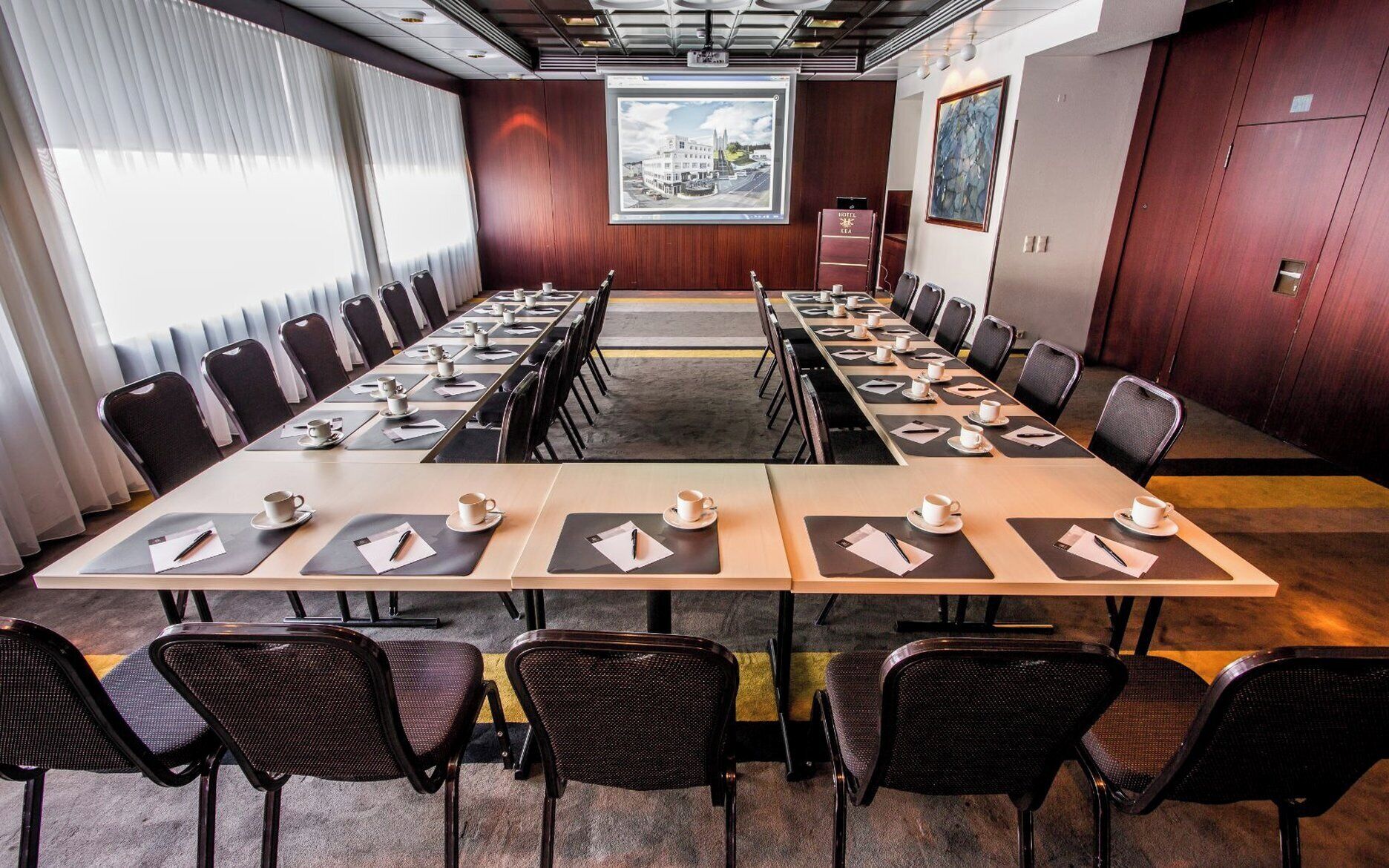 A conference room with long tables, chairs, and a large screen at the front.
