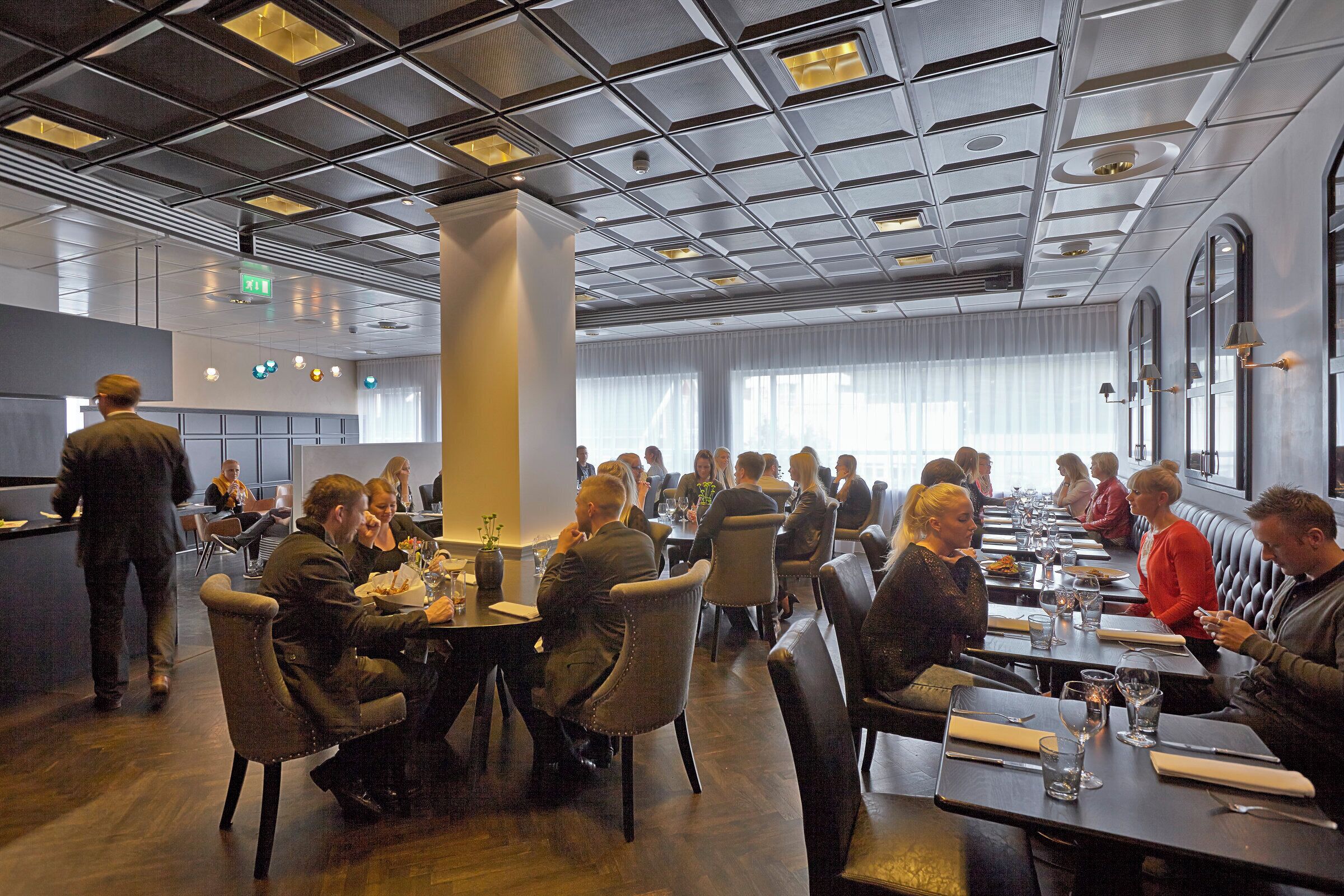 A spacious restaurant with high ceilings and modern lighting fixtures. Tables are arranged in a casual layout, and there is a bar area visible in the background.
