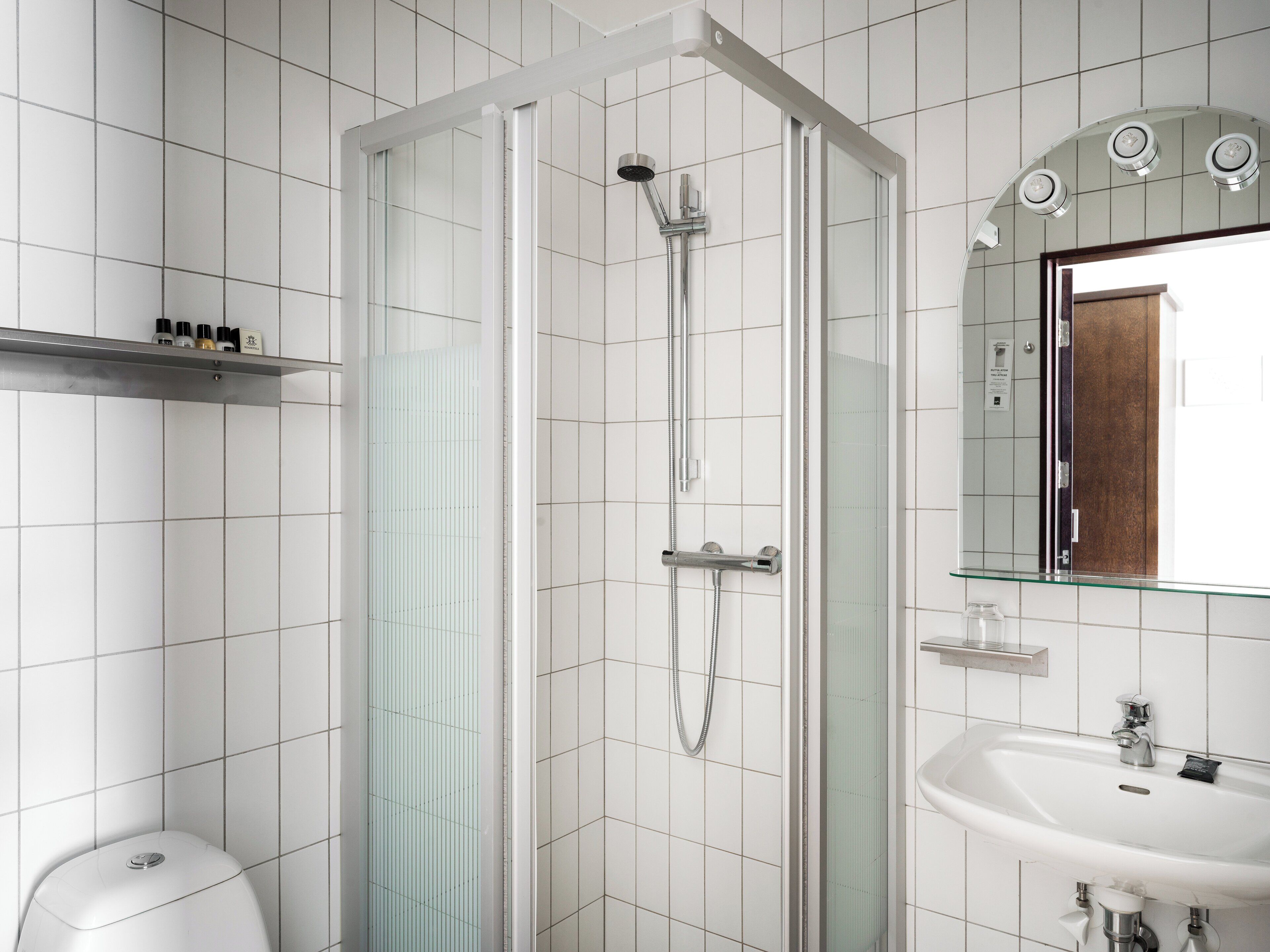 Ein modernes Badezimmer mit einer gläsernen Duschkabine, einem weißen Waschbecken und einem Spiegel, der eine Holztür reflektiert.