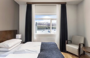 Habitación de hotel con cama, una silla, una mesita, ventana con cortinas y vista a un paisaje.