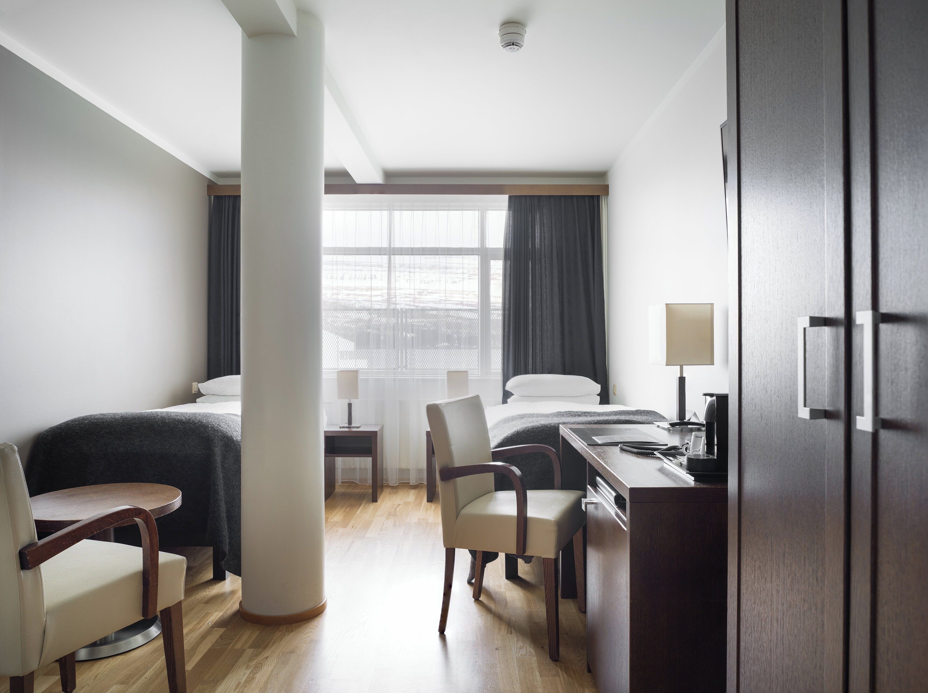 Habitación de hotel con dos camas, un escritorio, una silla, una lámpara y vistas a la ciudad desde la ventana.