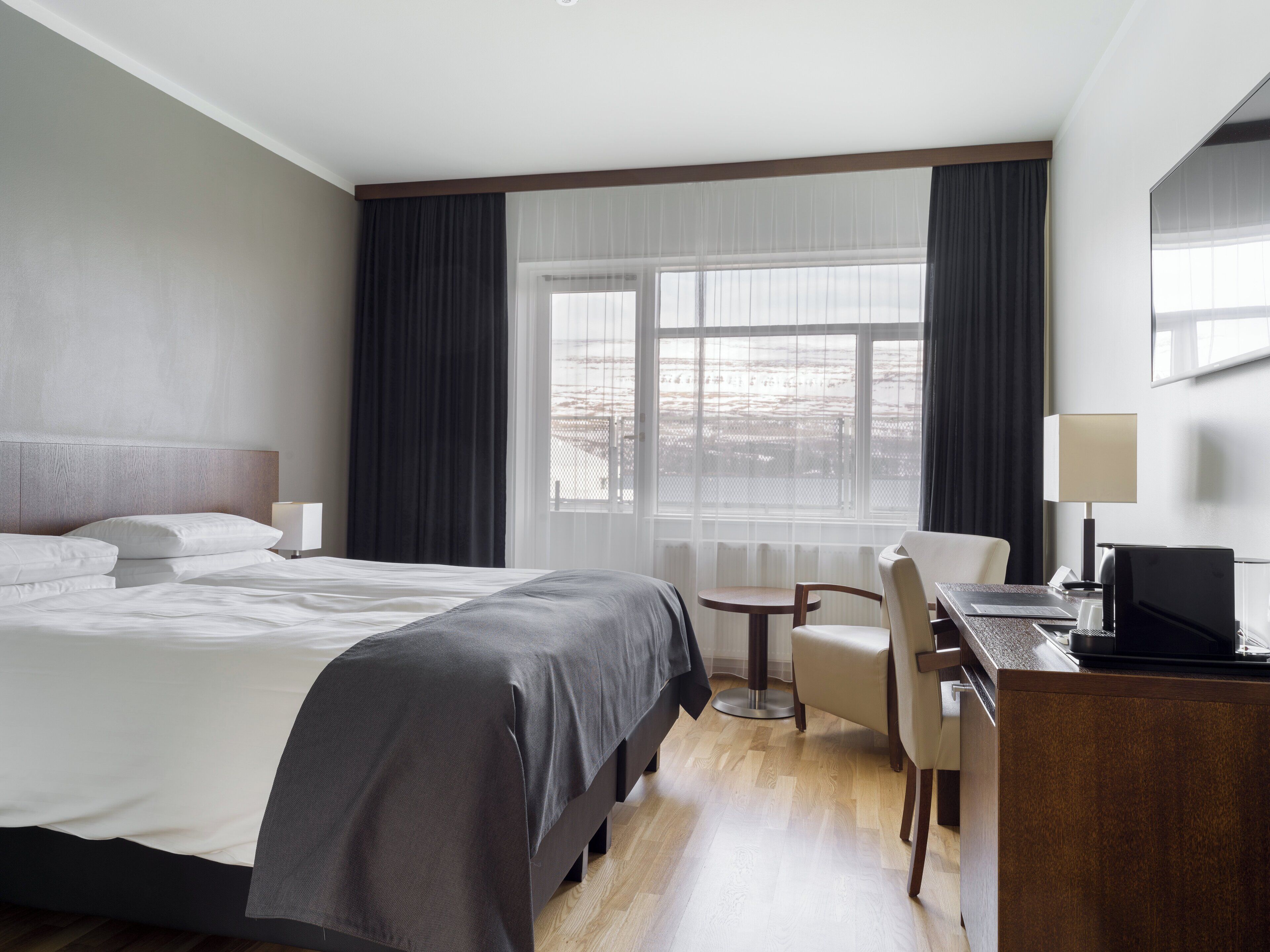 A hotel room with a large bed, a desk with a chair, a small table, and a window with curtains.