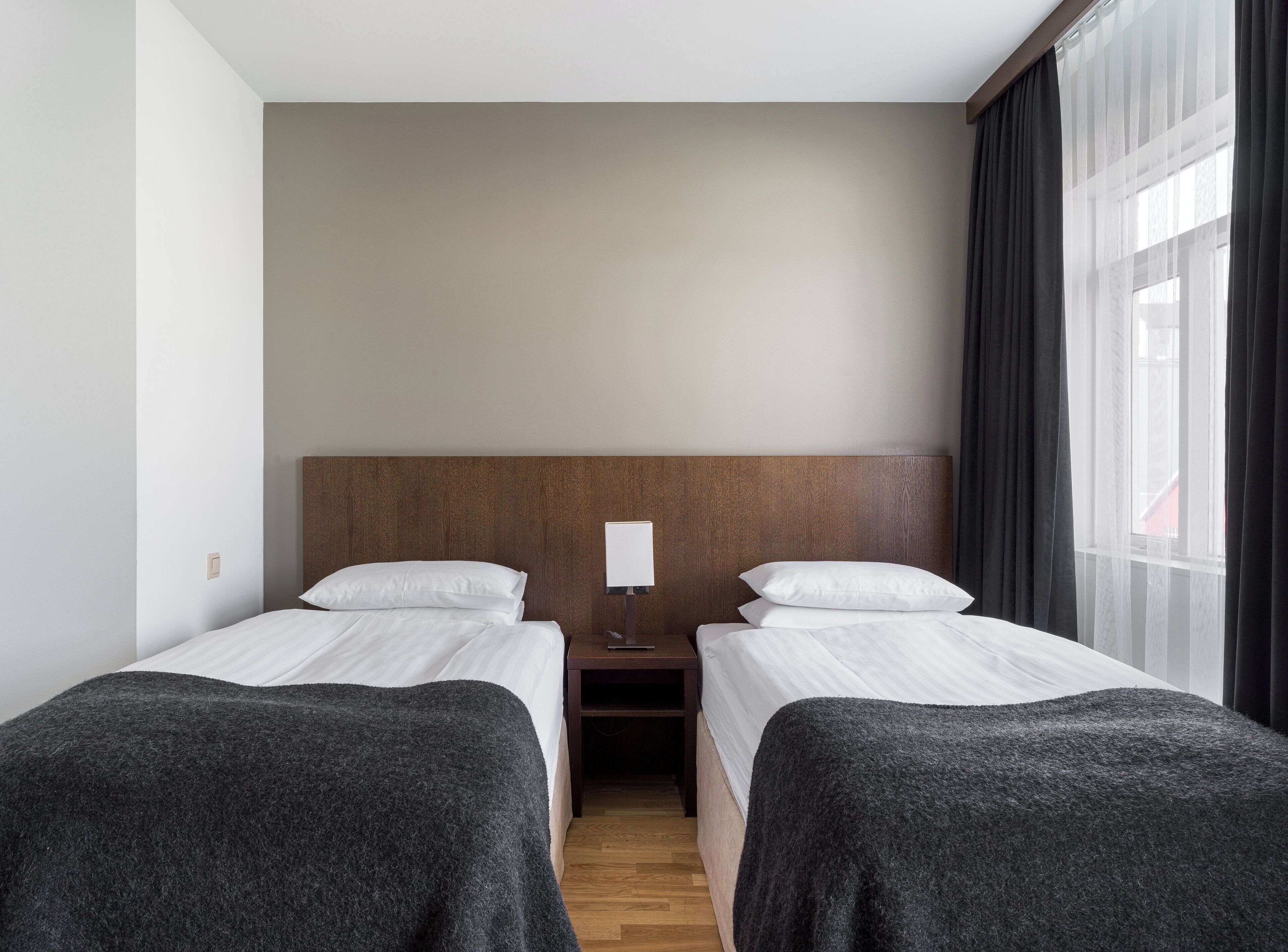 Ein Hotelzimmer mit zwei Betten, einem Holzkopfteil, einem Nachttisch mit Lampe und einem Fenster mit Vorhängen.