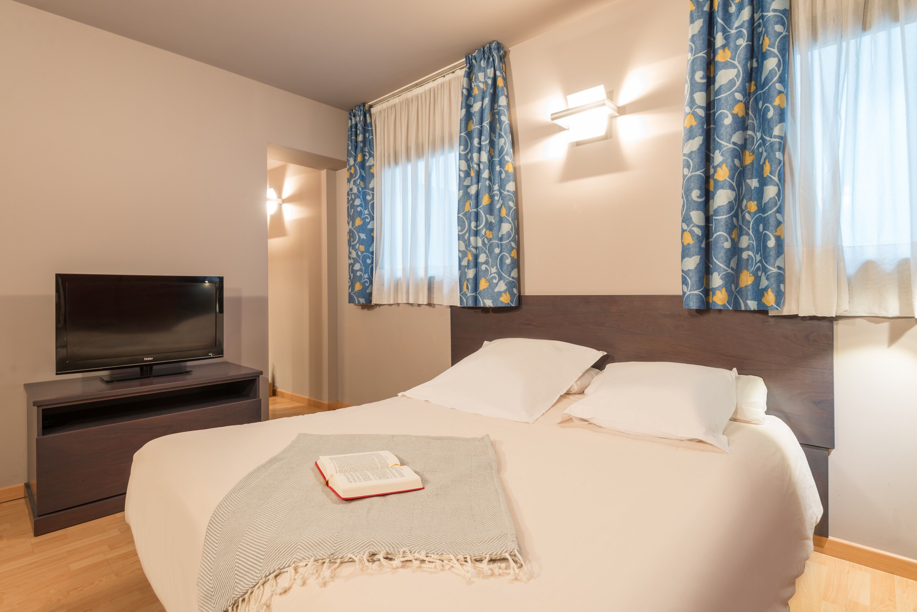 A neatly arranged hotel room with a bed, a TV on a stand, and curtains with a floral pattern.