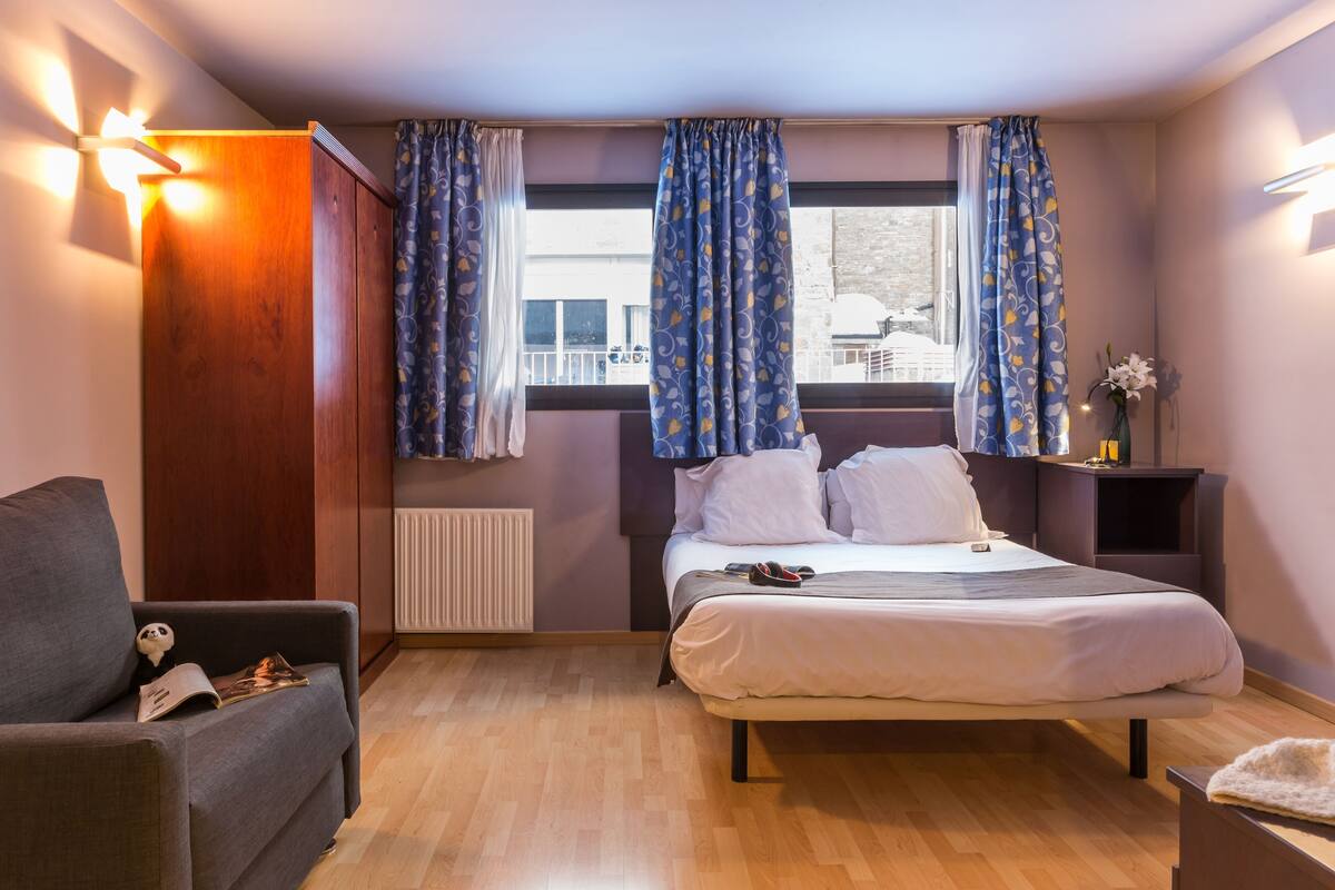 A bedroom with a bed, a sofa, a nightstand, and a window with curtains.