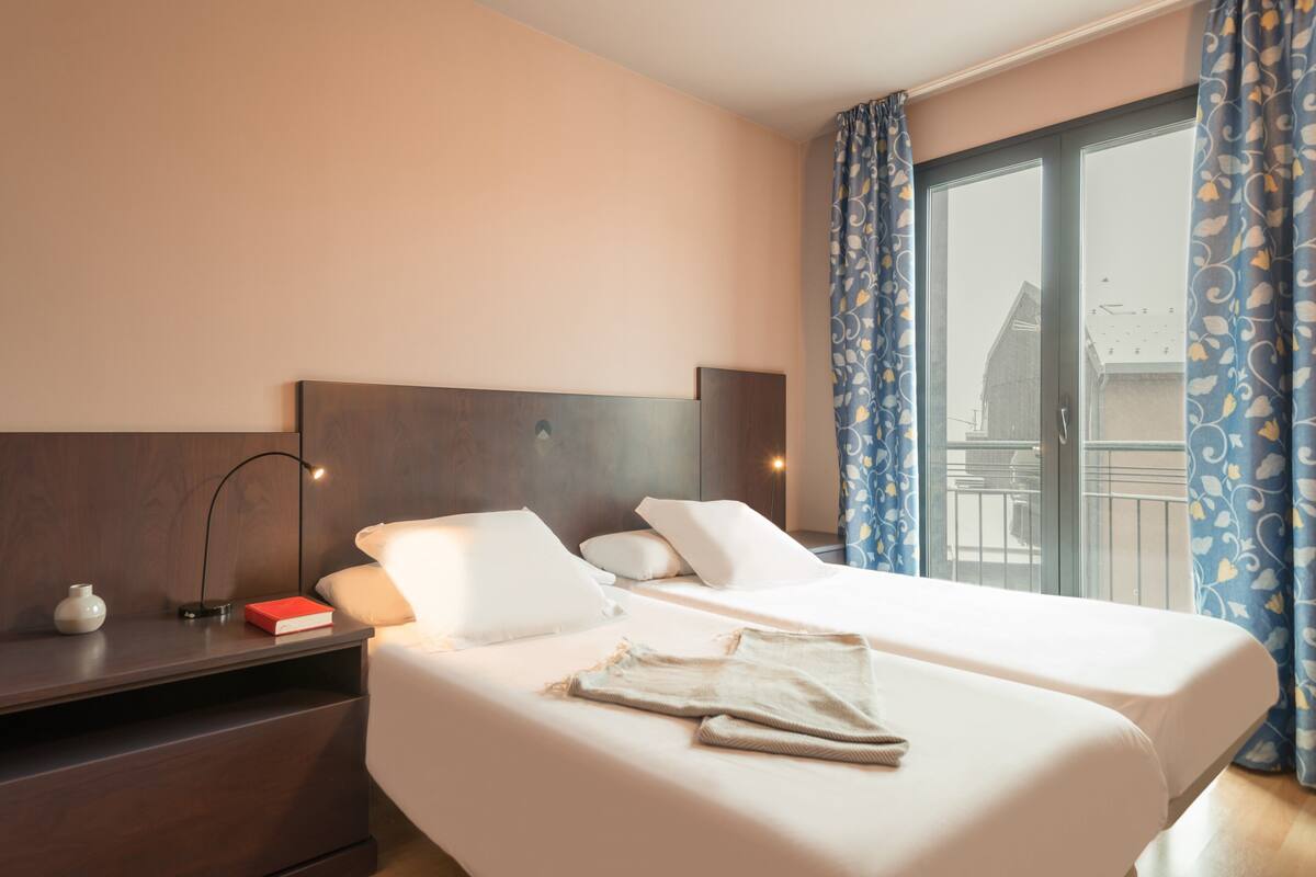 A hotel room with a large bed, a bedside table with a lamp, a small vase, and a red book.
