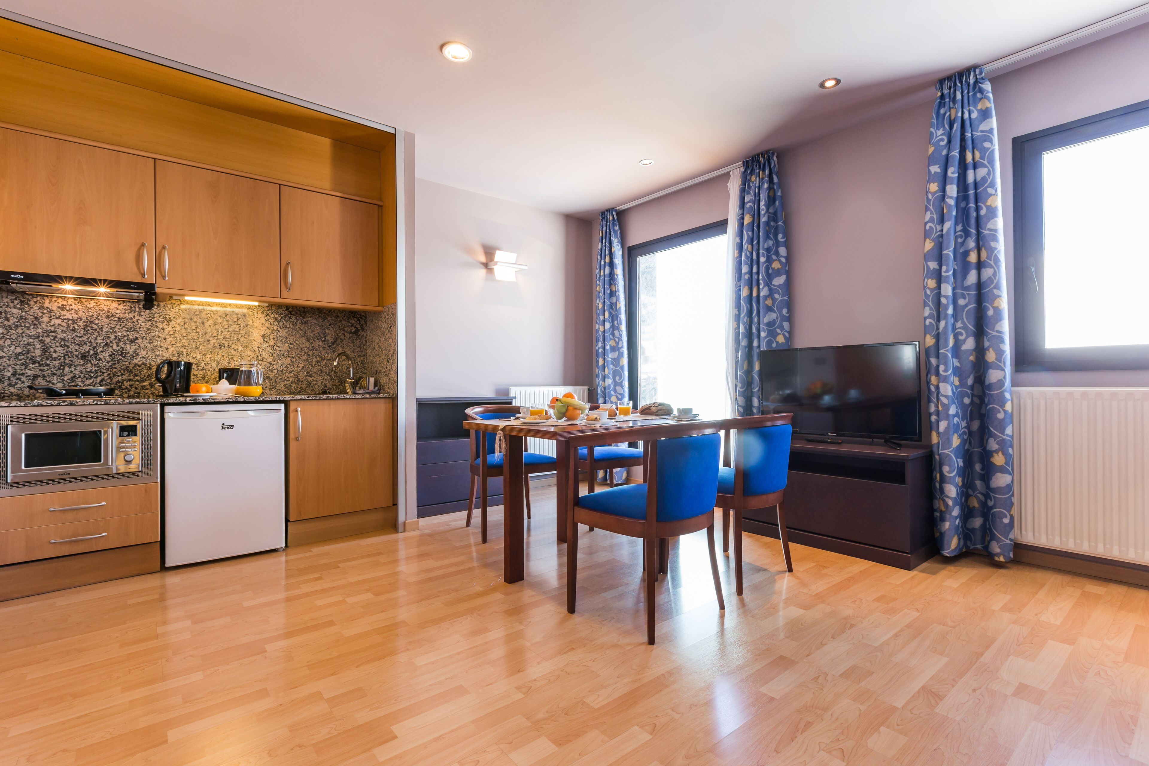 Una cocina moderna con armarios de madera, un microondas empotrado y un refrigerador pequeño. Hay una zona de comedor con una mesa y sillas, y un televisor de pantalla plana montado en la pared.