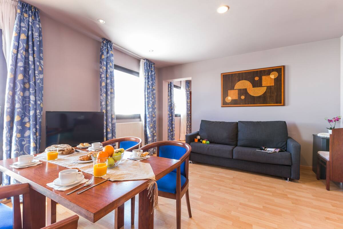 A dining area with a wooden table set for breakfast, a sofa, and a TV in the background.