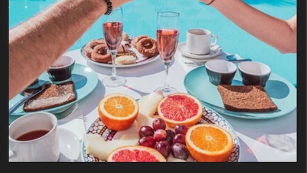 A poolside breakfast setting with a table set for two, featuring orange juice, pastries, and a variety of fruits.