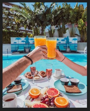 Desayuno junto a la piscina con una mesa para dos, que incluye zumo de naranja, bollería y diversas frutas.
