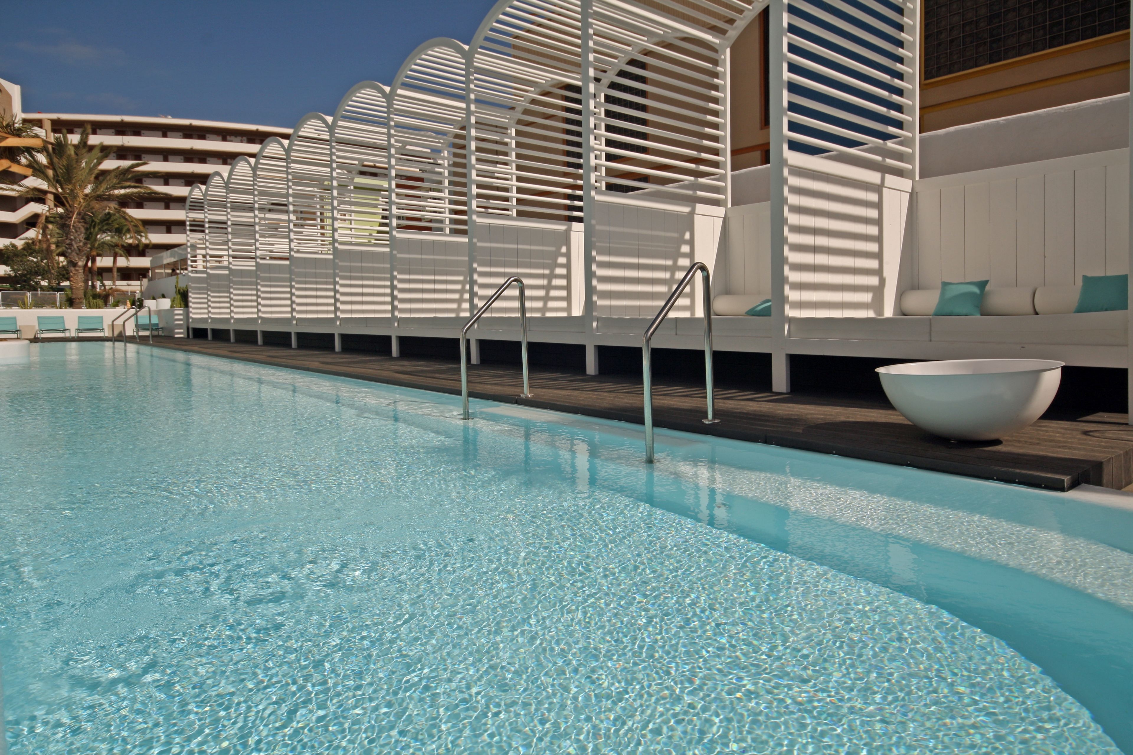 A swimming pool with clear water, surrounded by white lounge chairs and a white wall with a curved design.