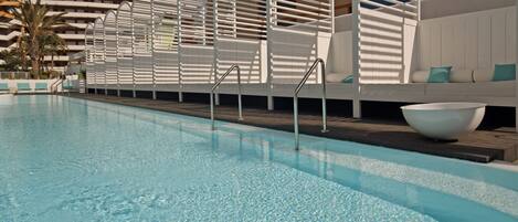 A swimming pool with clear water, surrounded by white lounge chairs and a white wall with a curved design.