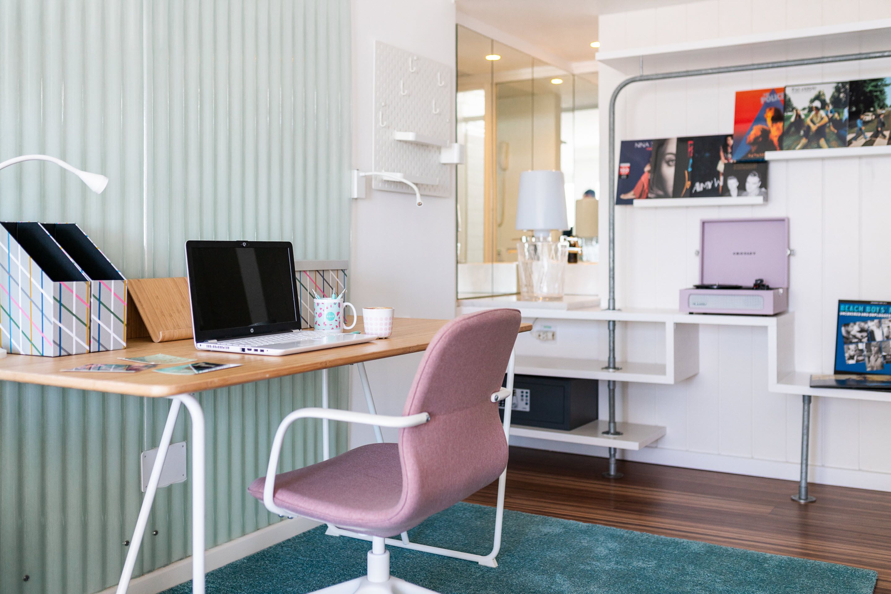 Ein modernes Hotelzimmer mit Schreibtisch aus Holz, einem pinken Stuhl, einem Laptop, einer Kaffeetasse und einem Bücherregal.