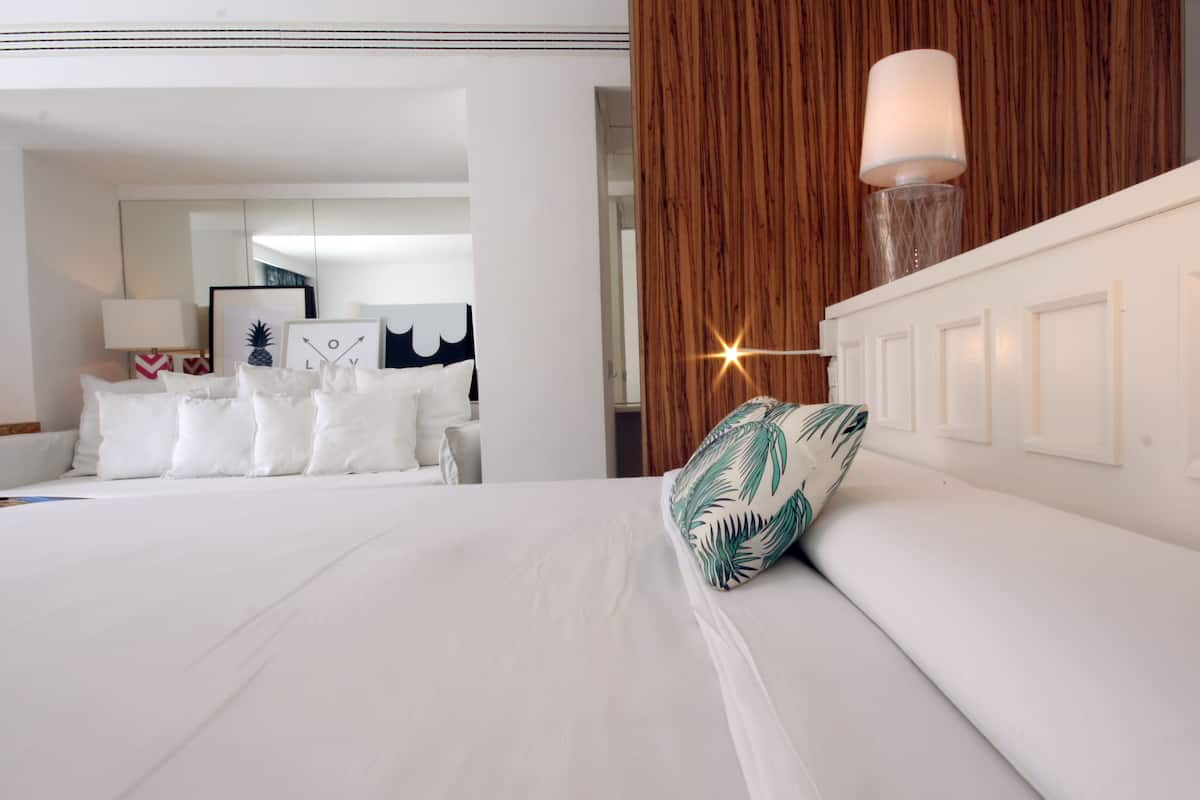 Ein Hotelzimmer mit einem großen Bett, weißer Bettwäsche, einem Kissen mit tropischem Blattmuster, einer Wand mit Holzvertäfelung und einem Nachttisch mit Lampe.
