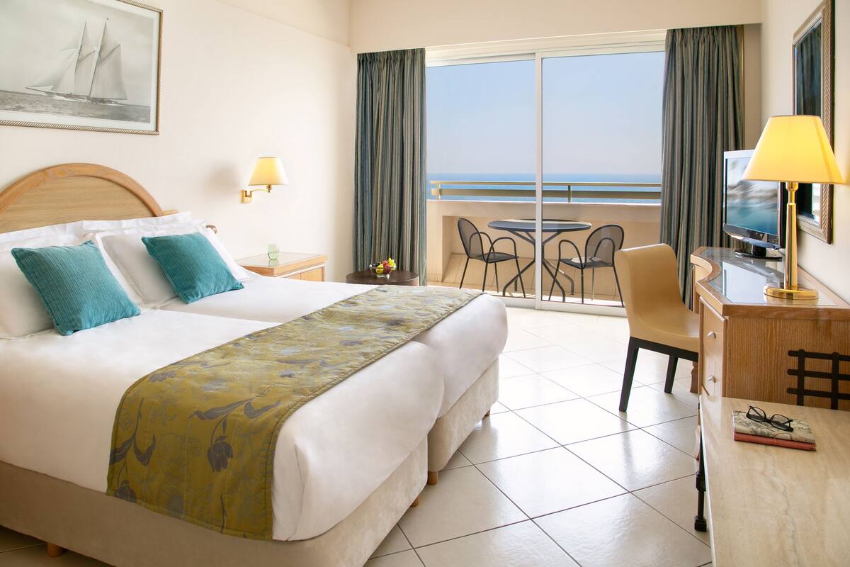 Quarto de hotel com uma cama grande, uma escrivaninha com abajur, televisão e varanda com vista para o mar.