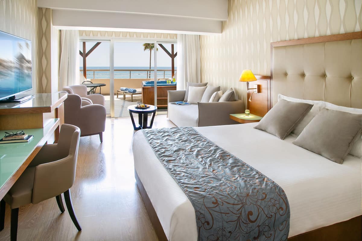 Quarto de hotel com uma cama grande, uma mesa com televisão, um sofá e varanda com vista para o oceano.