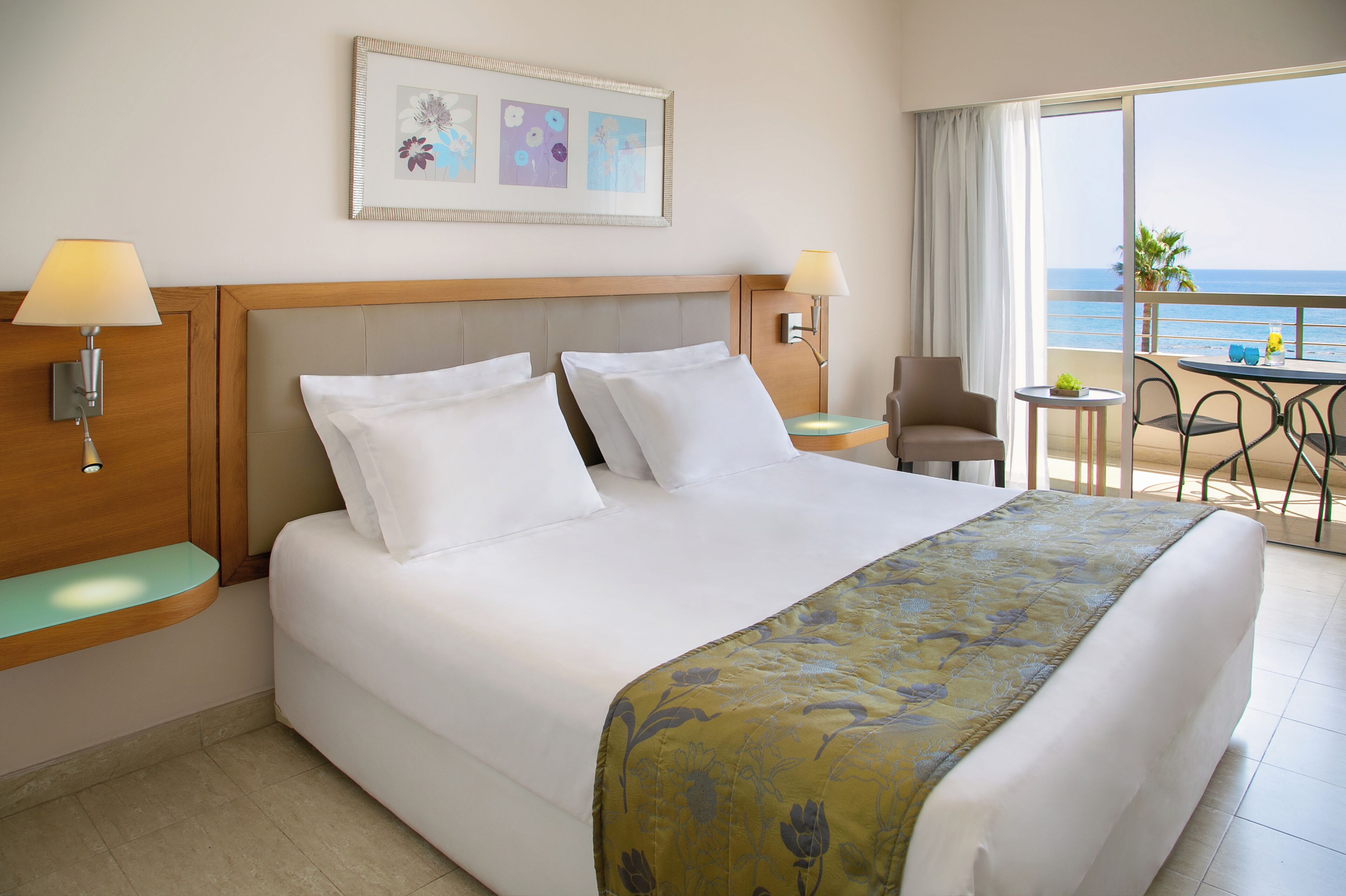 A hotel room with a bed, two bedside lamps, a small table with chairs, and a balcony with a view of the sea.